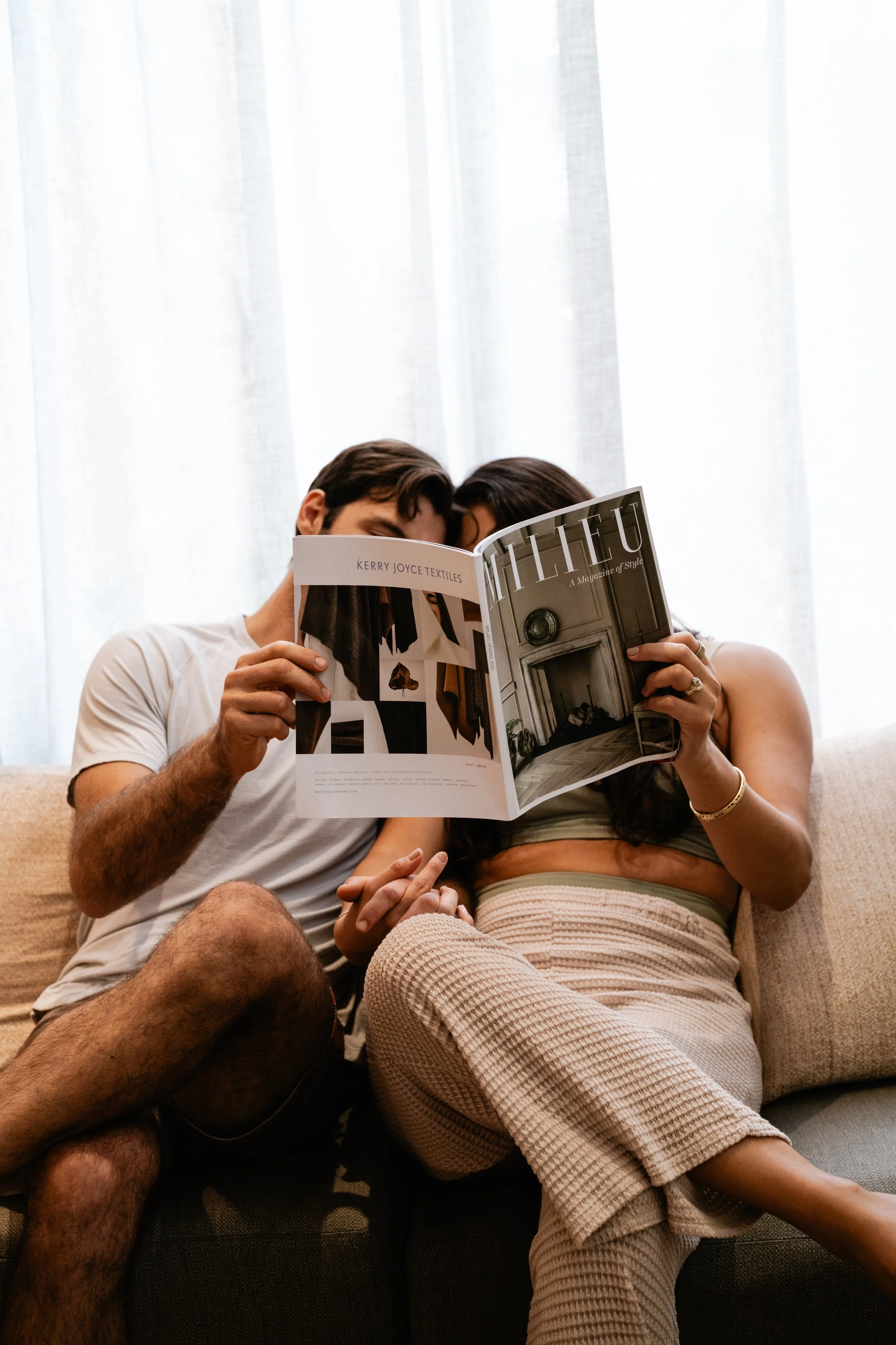A couple sitting on a beige couch, reading a magazine together and holding hands, in front of a light-colored curtain.