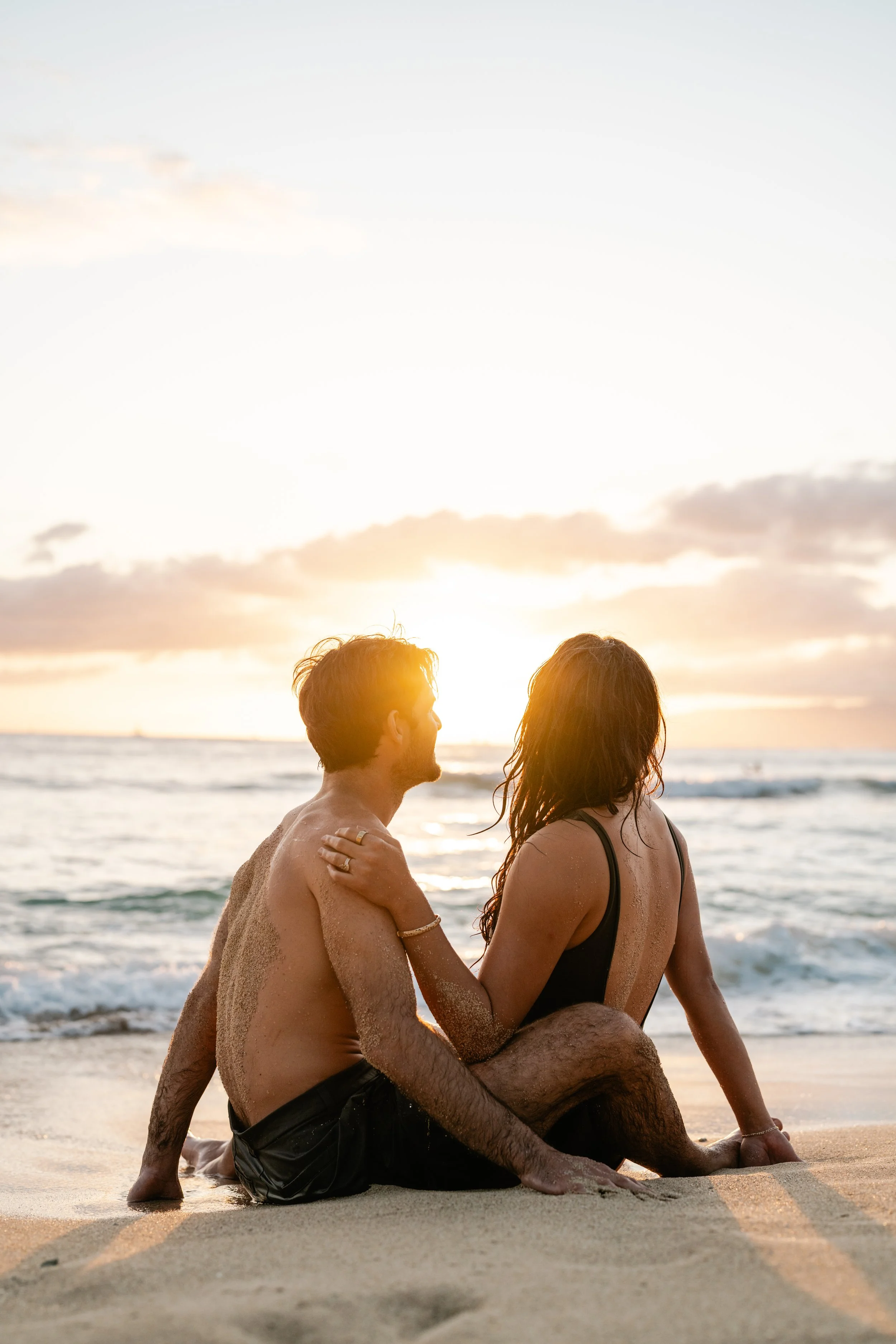 A couple sitting on the beach at sunset, facing the ocean, with the sun setting behind them.