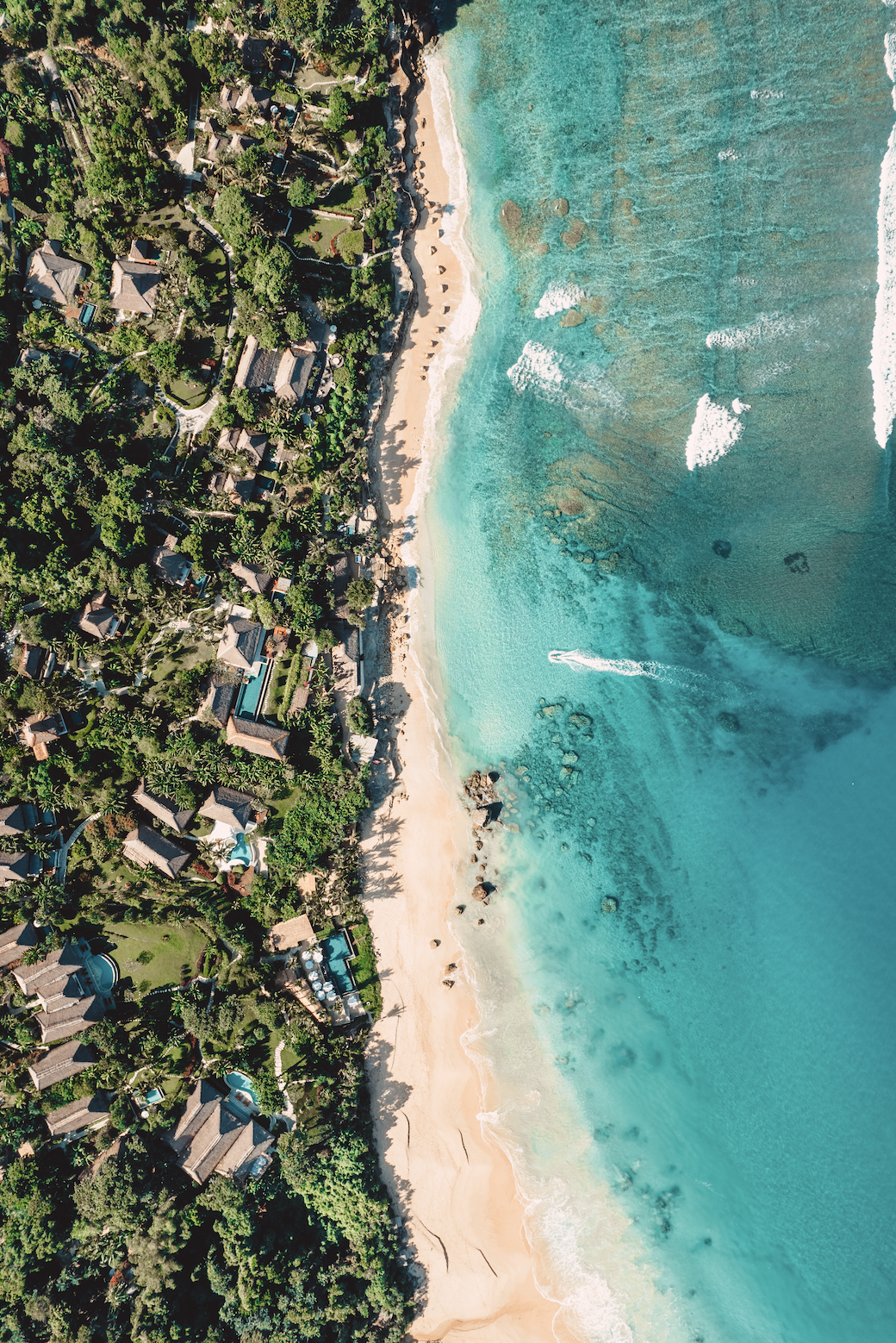 An aerial view of a tropical beach with turquoise waters, white sandy shoreline, and lush green vegetation with scattered houses nearby.