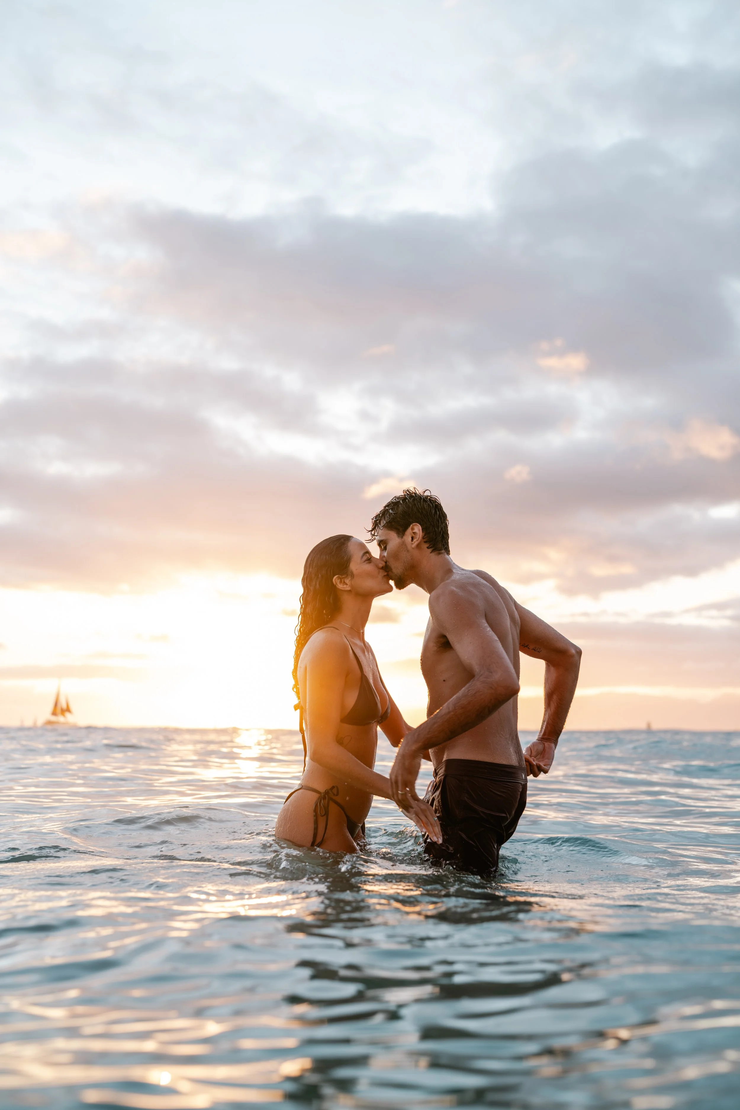 A couple in swimsuits sharing a kiss in the ocean at sunset.