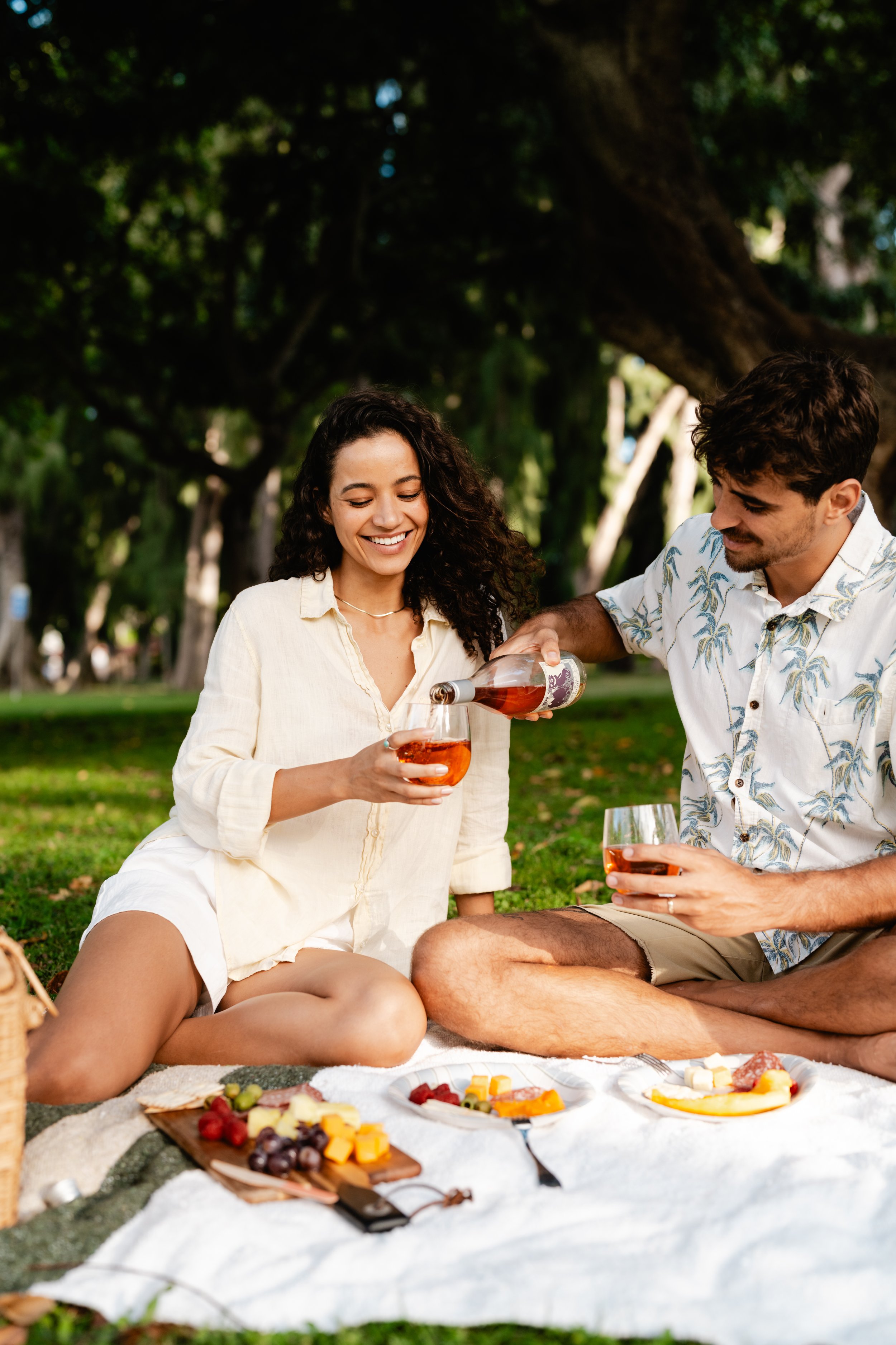A woman and a man having a picnic outdoors, pouring wine into glasses, with a picnic spread of cheese, grapes, and crackers on a blanket