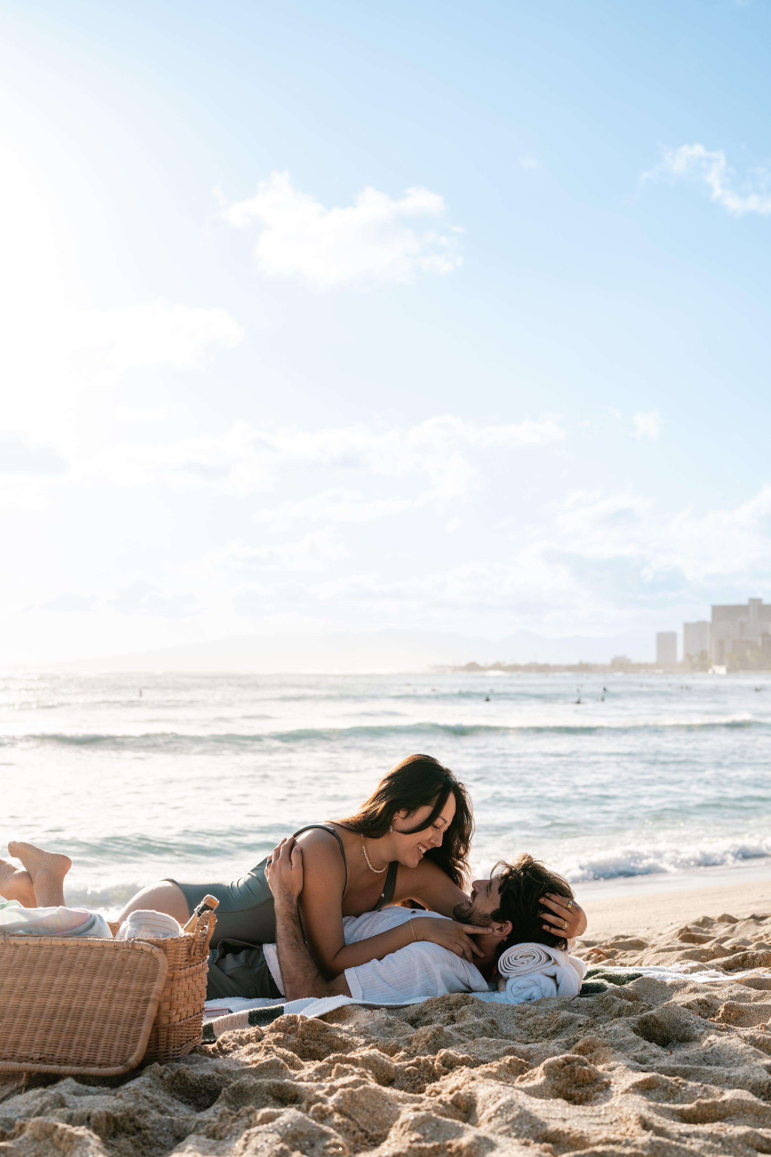 A woman and a man relaxing and playing on the sandy beach near the ocean, with buildings visible in the distance, under a bright blue sky with some clouds.