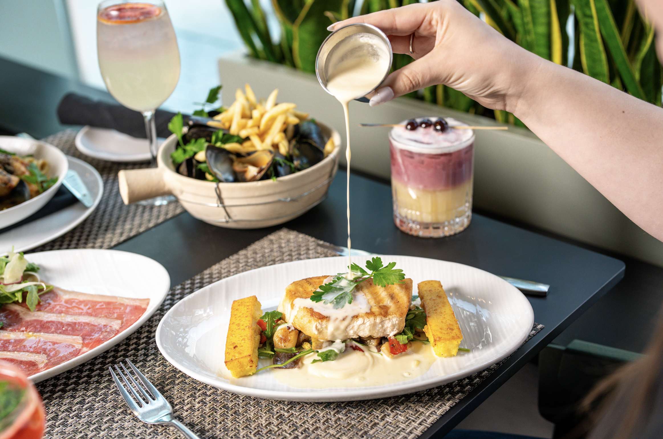 A hand pouring a creamy sauce over a plated dish of fish with vegetables and two yellow sides, at a table with salads, a bowl of mussels with fries, and layered cocktails in the background.