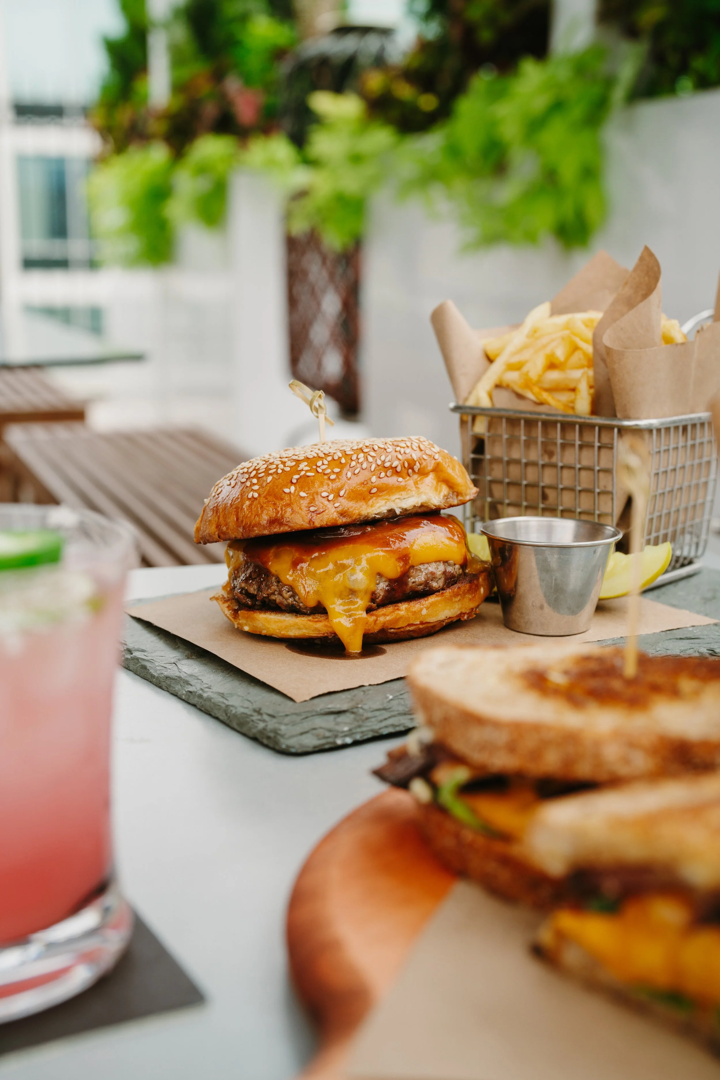 Cheeseburger with melted cheese on a sesame seed bun, served on a slate plate, with a side of fries in a metal basket and a lemon wedge, on an outdoor table.