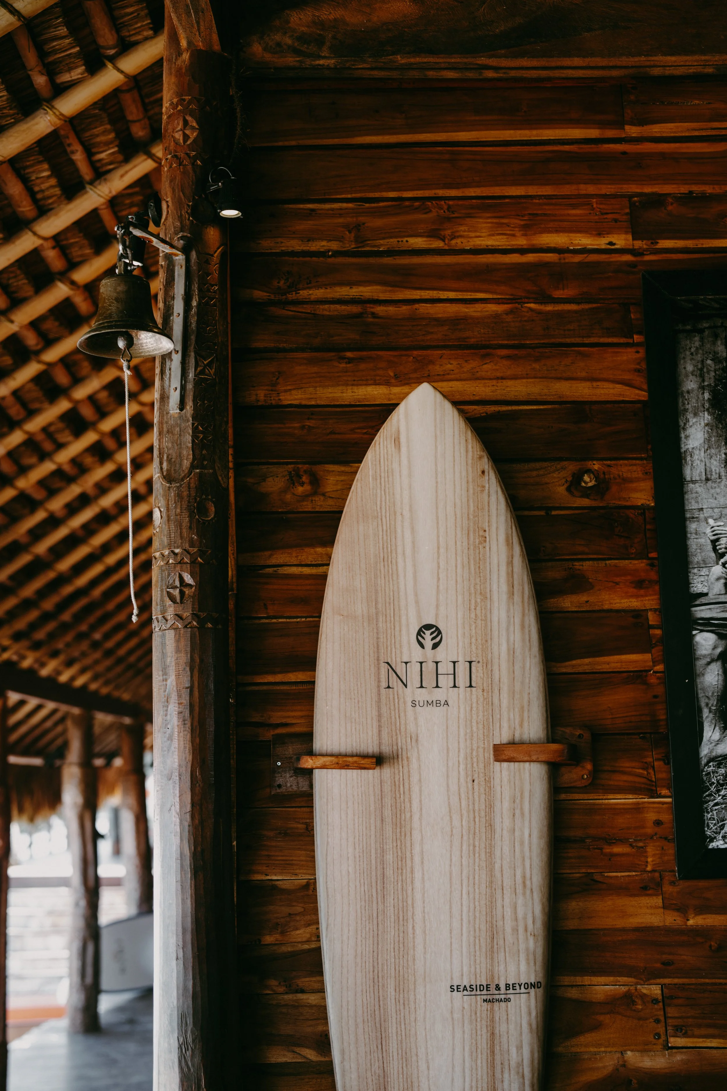 A wooden surfboard with the words 'NIHI SUMBA' and 'SEASIDE & BEYOND' leaning against a wooden wall in a rustic, beach-themed interior.
