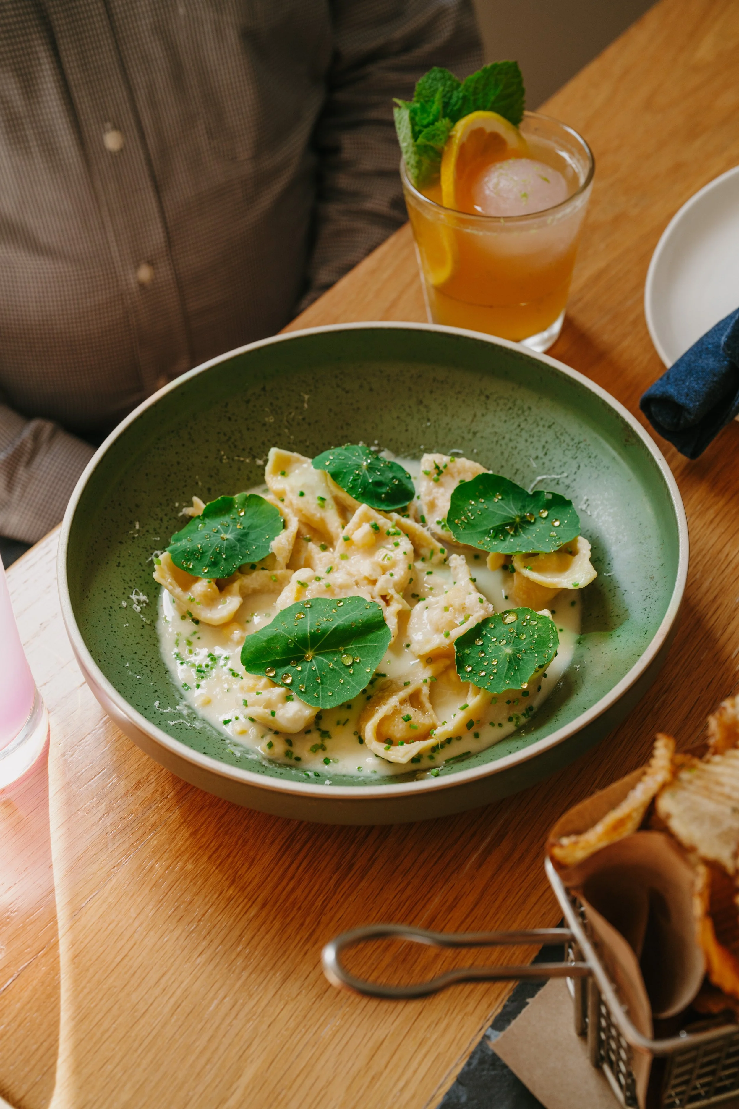 A bowl of pasta with green leaves and herbs, served with a creamy white sauce. A drink with lemon, orange slices, mint leaves, and ice is on the table.