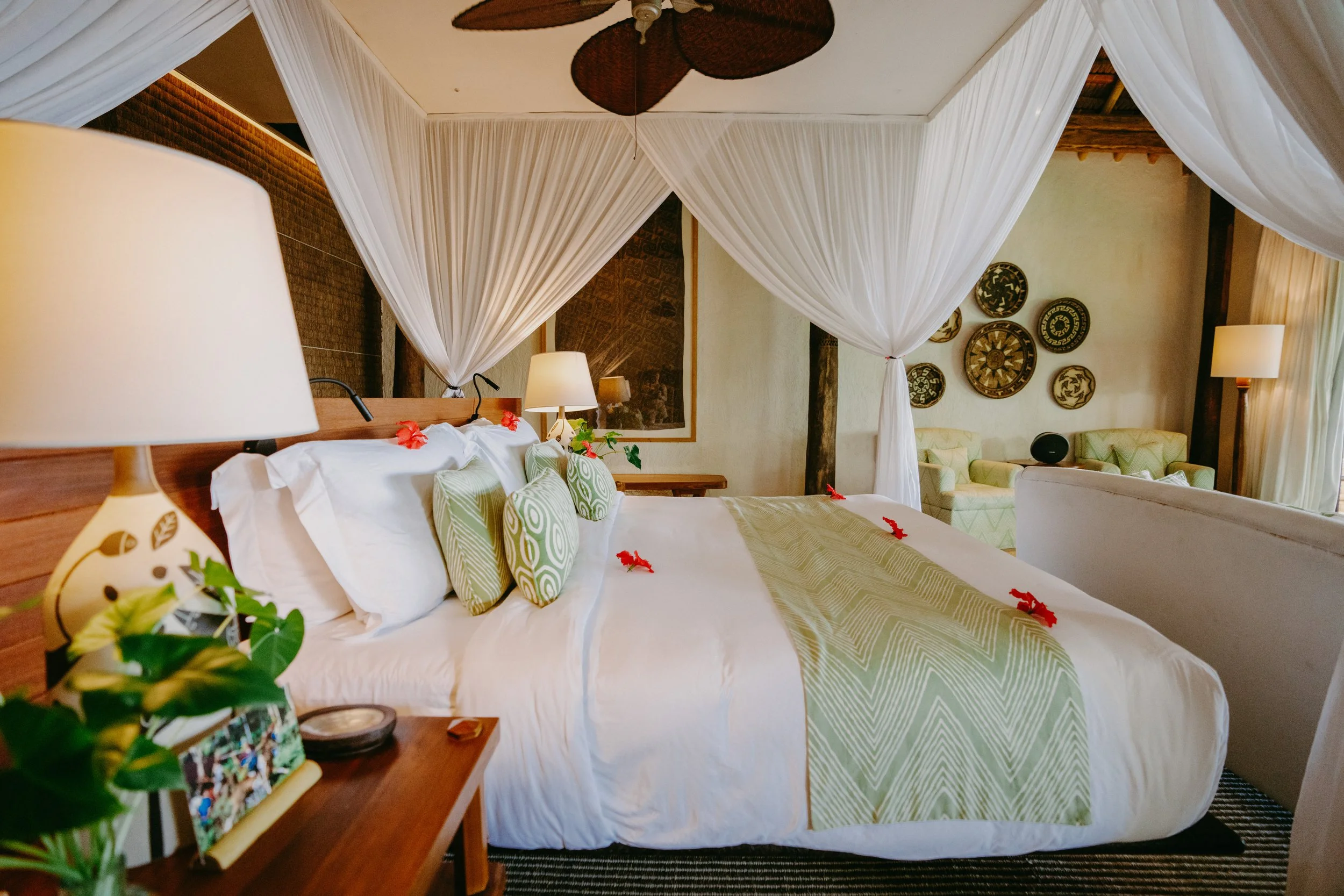 A tropical-style bedroom with a large bed decorated with green and white pillows, white linens, and a green bed runner, surrounded by white curtains and tropical decor including a ceiling fan and wall art.