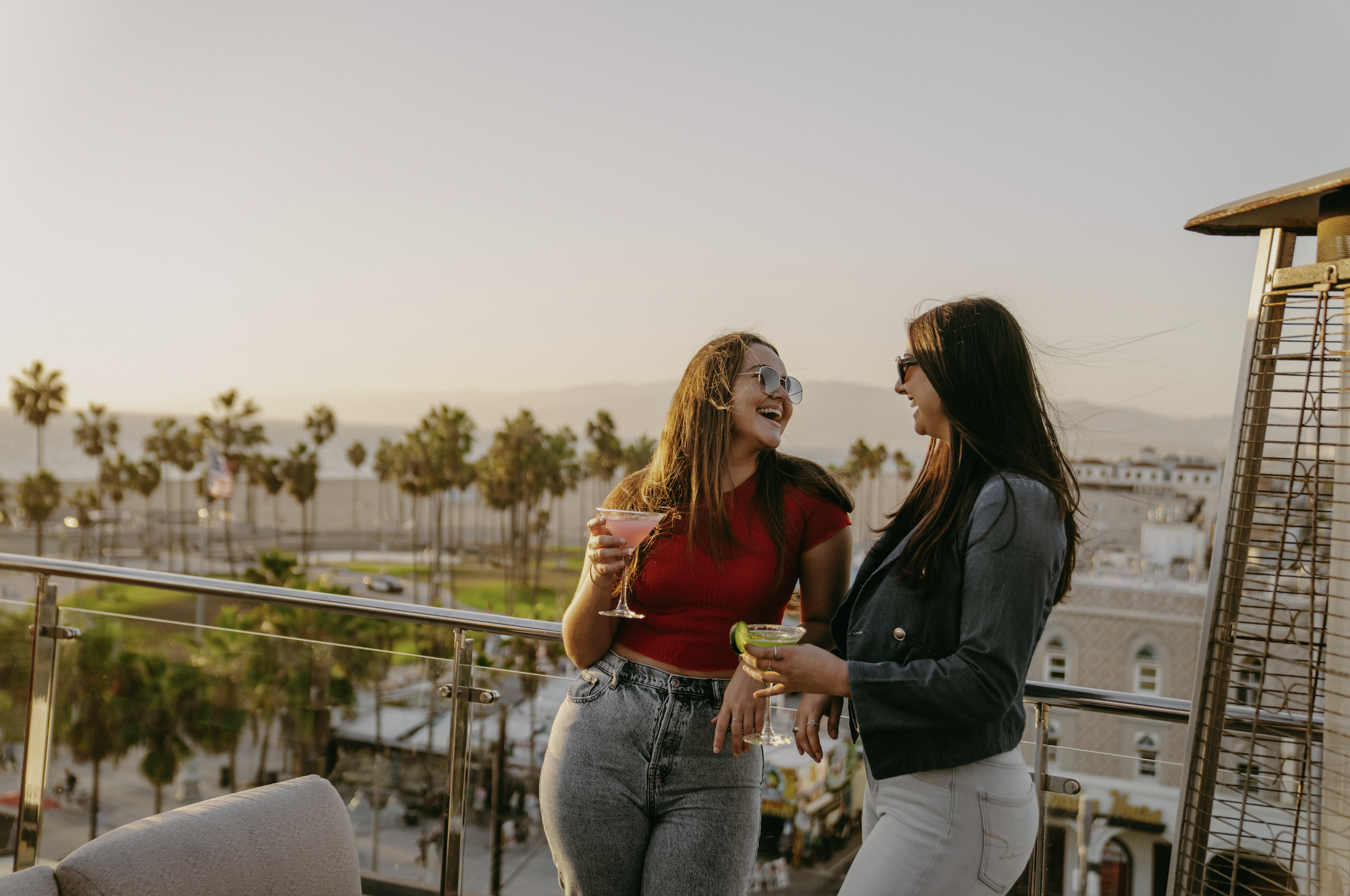Two women laughing and talking on a rooftop with drinks during sunset, with palm trees and buildings in the background.