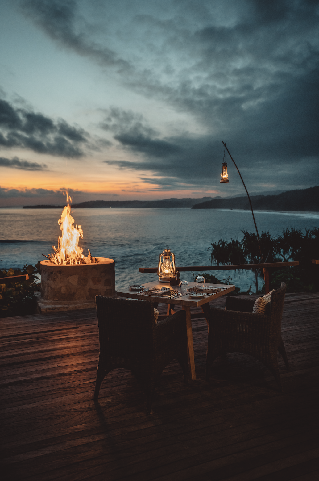 A cozy outdoor dining setup on a wooden deck overlooking the ocean at sunset, with a fire pit, a table set with glasses and plates, and lanterns providing warm lighting.