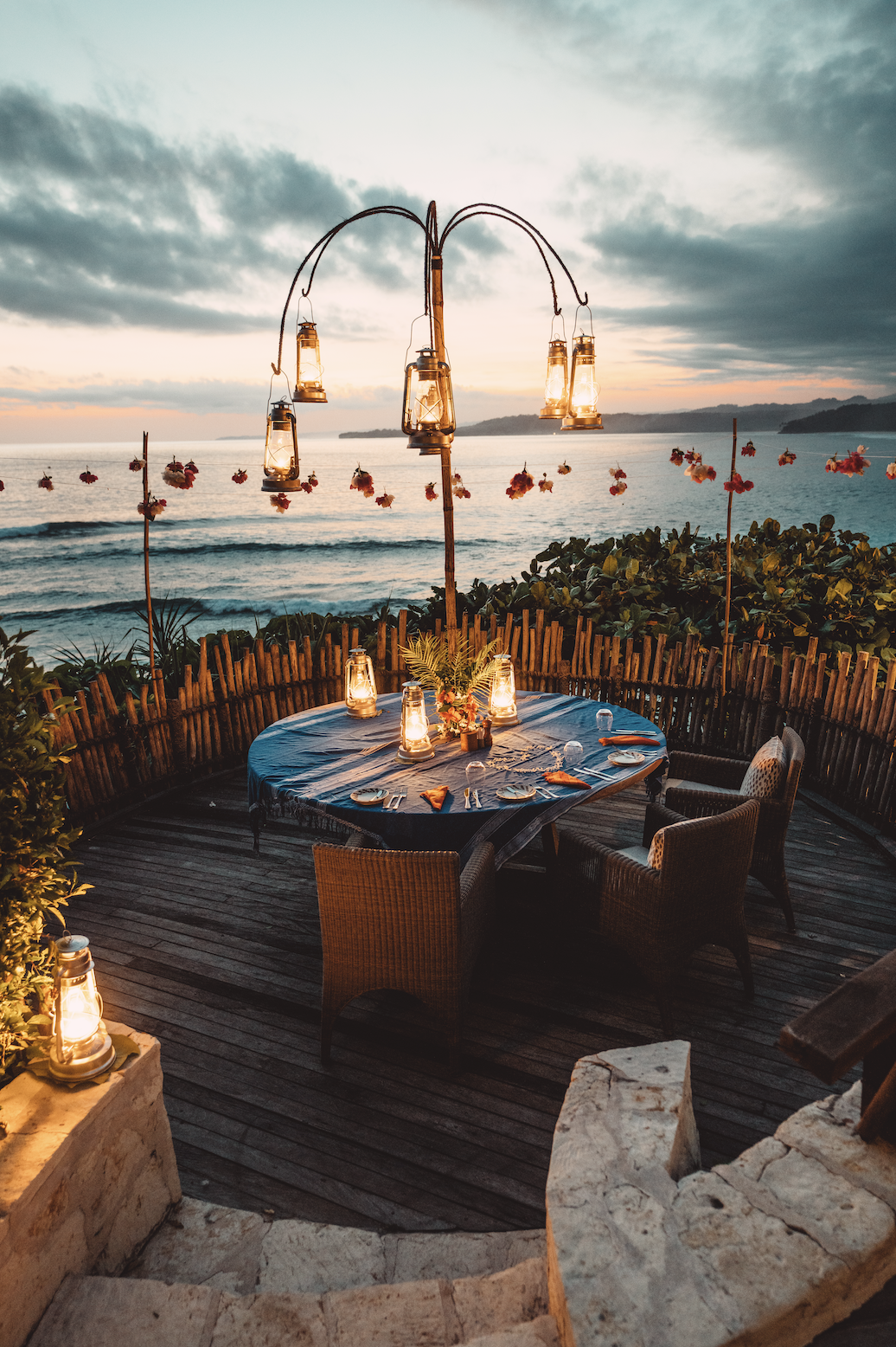 Outdoor dinner setup on a wooden deck overlooking the ocean at sunset, with a round table, wicker chairs, lanterns, and hanging lantern lights.