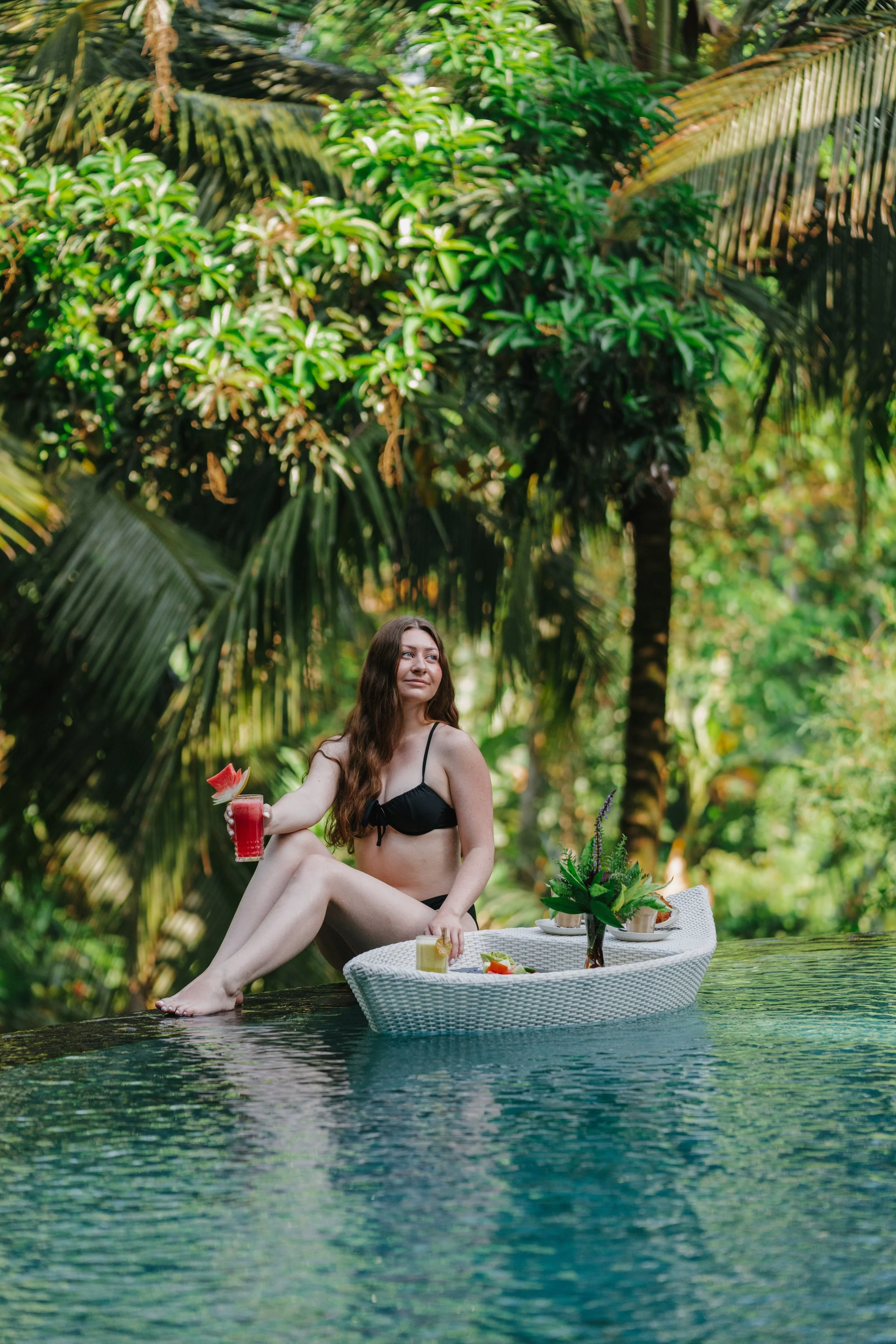 A woman in a black bikini sitting by a pool, holding a red cocktail with a watermelon slice, surrounded by lush tropical greenery and a decorative tray with flowers and drinks.