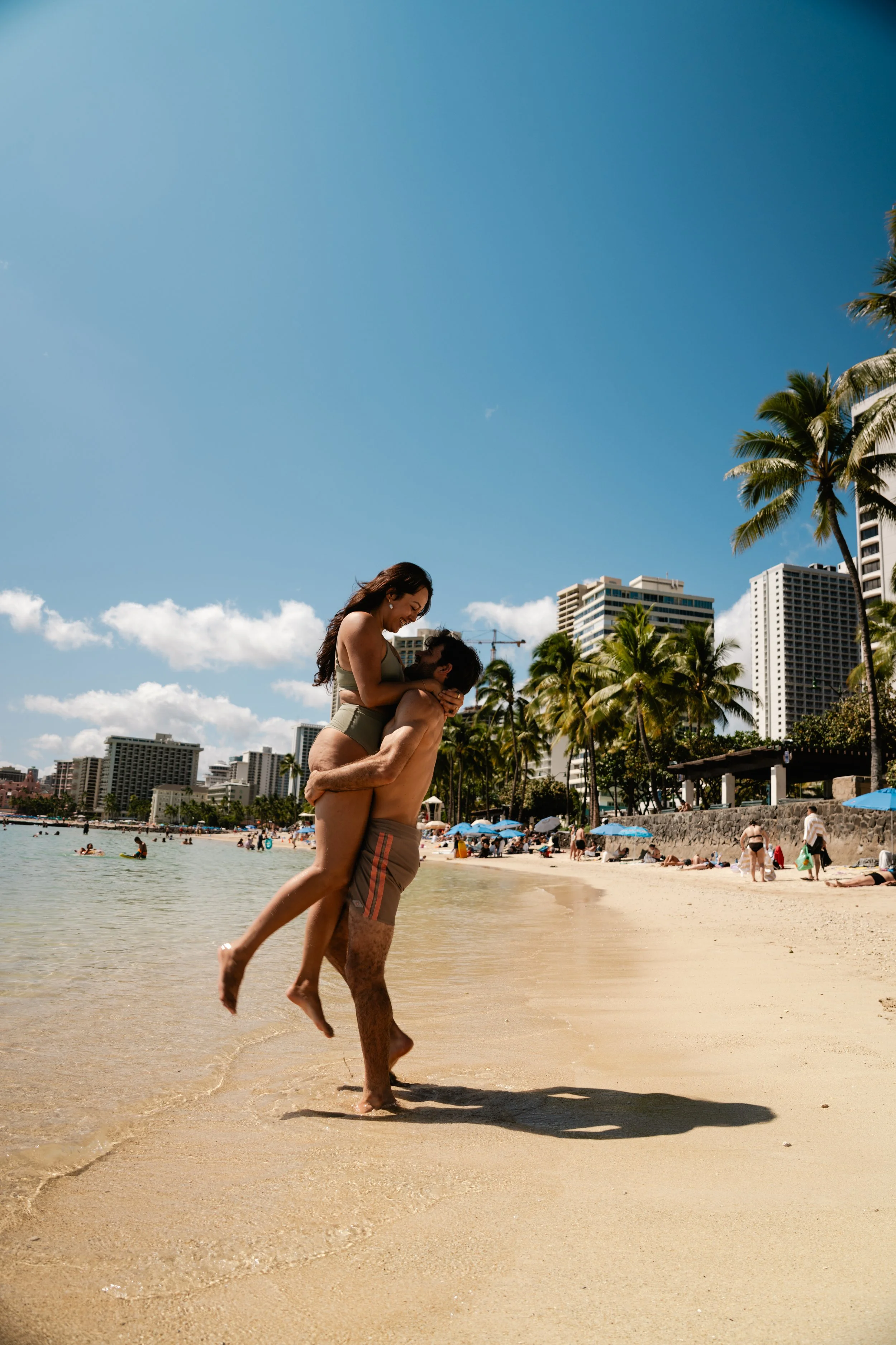 A couple enjoying a romantic moment on a sunny beach with palm trees, high-rise buildings, and umbrellas in the background.