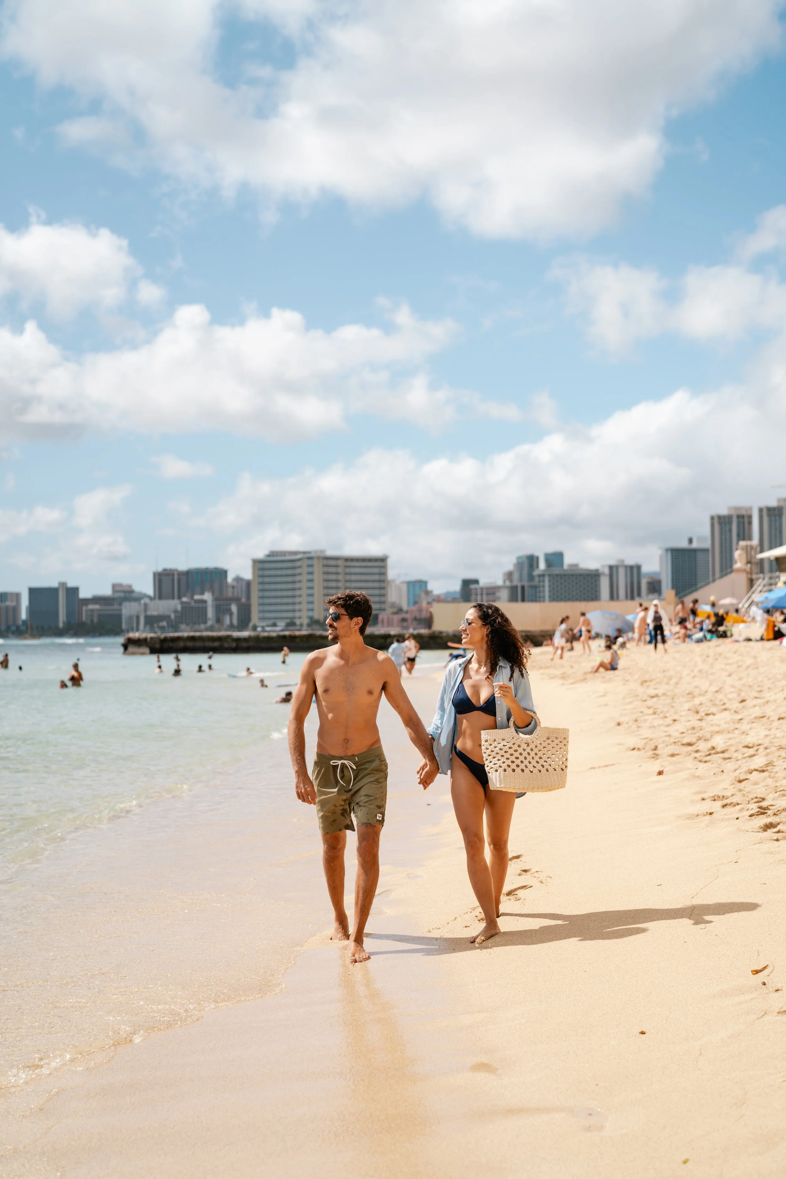 Hotel & Resort Photography & Videography. A couple walking hand in hand along a sunny beach with city skyline in the background, people swimming and relaxing on the sand.