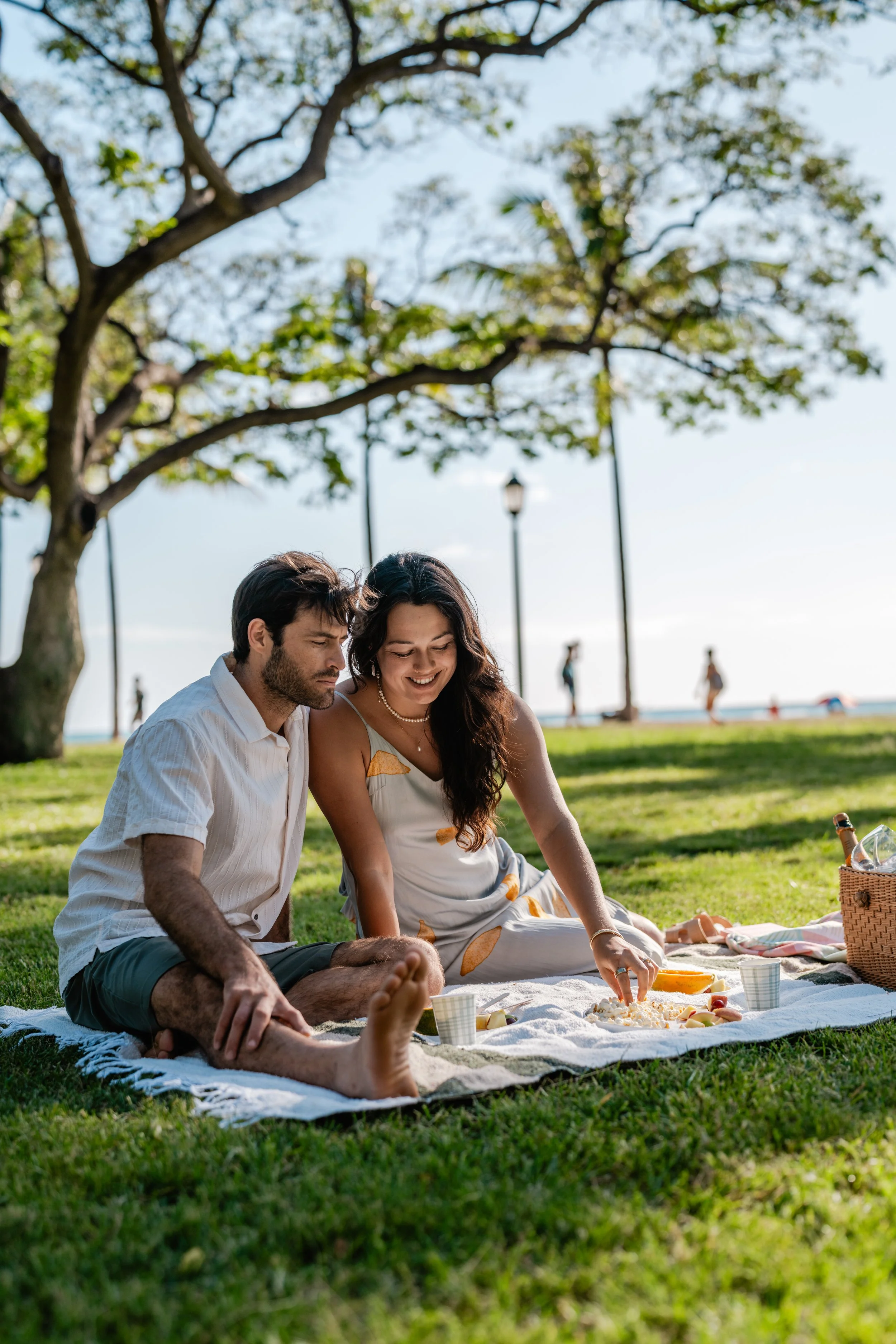 A couple having a picnic on a blanket in a park near the beach, with trees and a few people walking in the background.