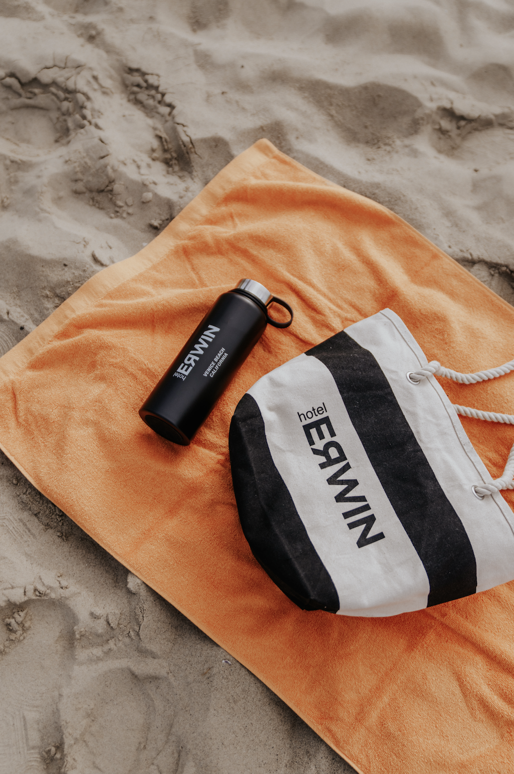 A black water bottle and a striped drawstring bag with the text "hotel EMELE" placed on an orange towel on sandy beach.