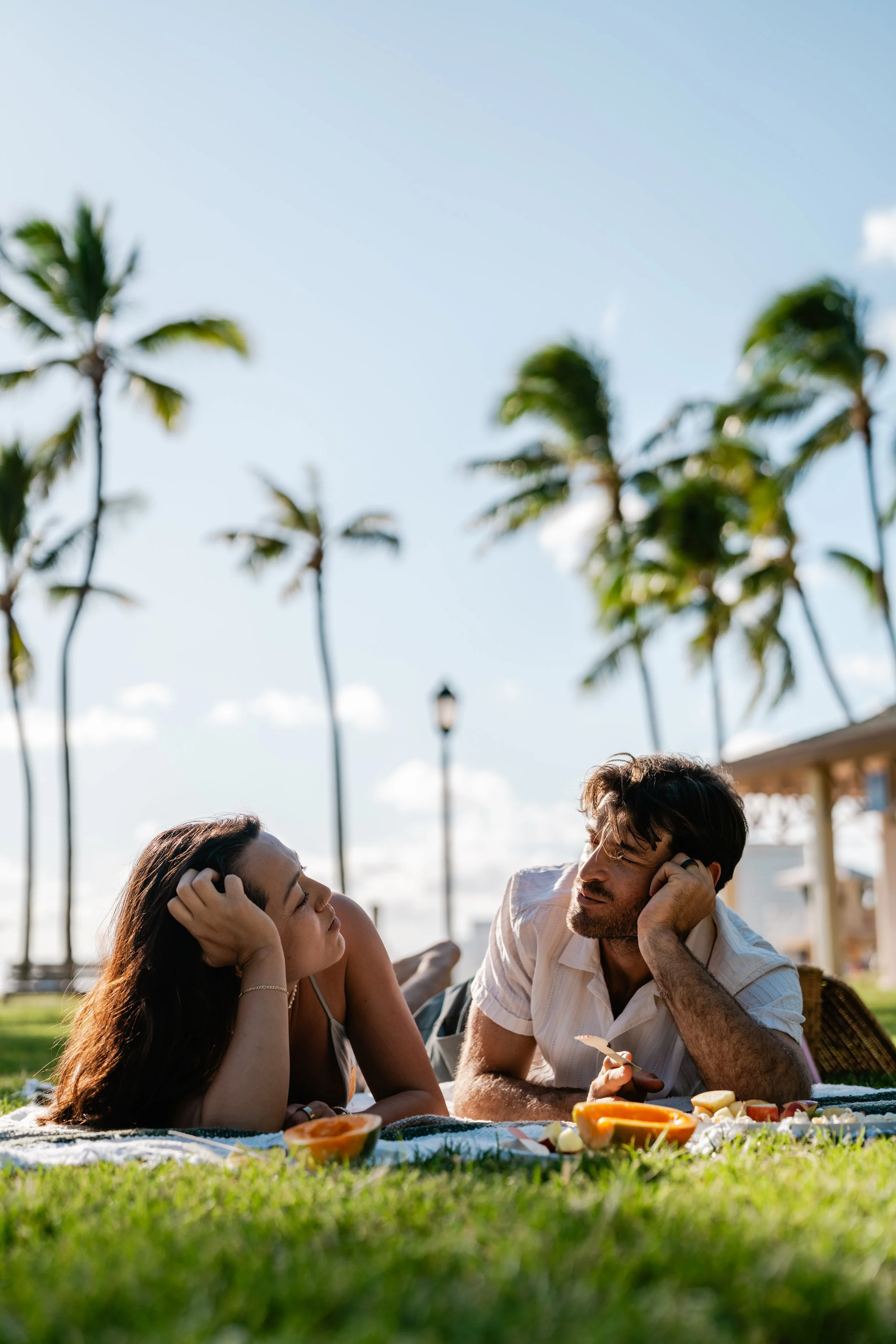 A man and woman lying on a blanket outdoors, having a picnic on a grassy area with palm trees and a clear blue sky in the background.