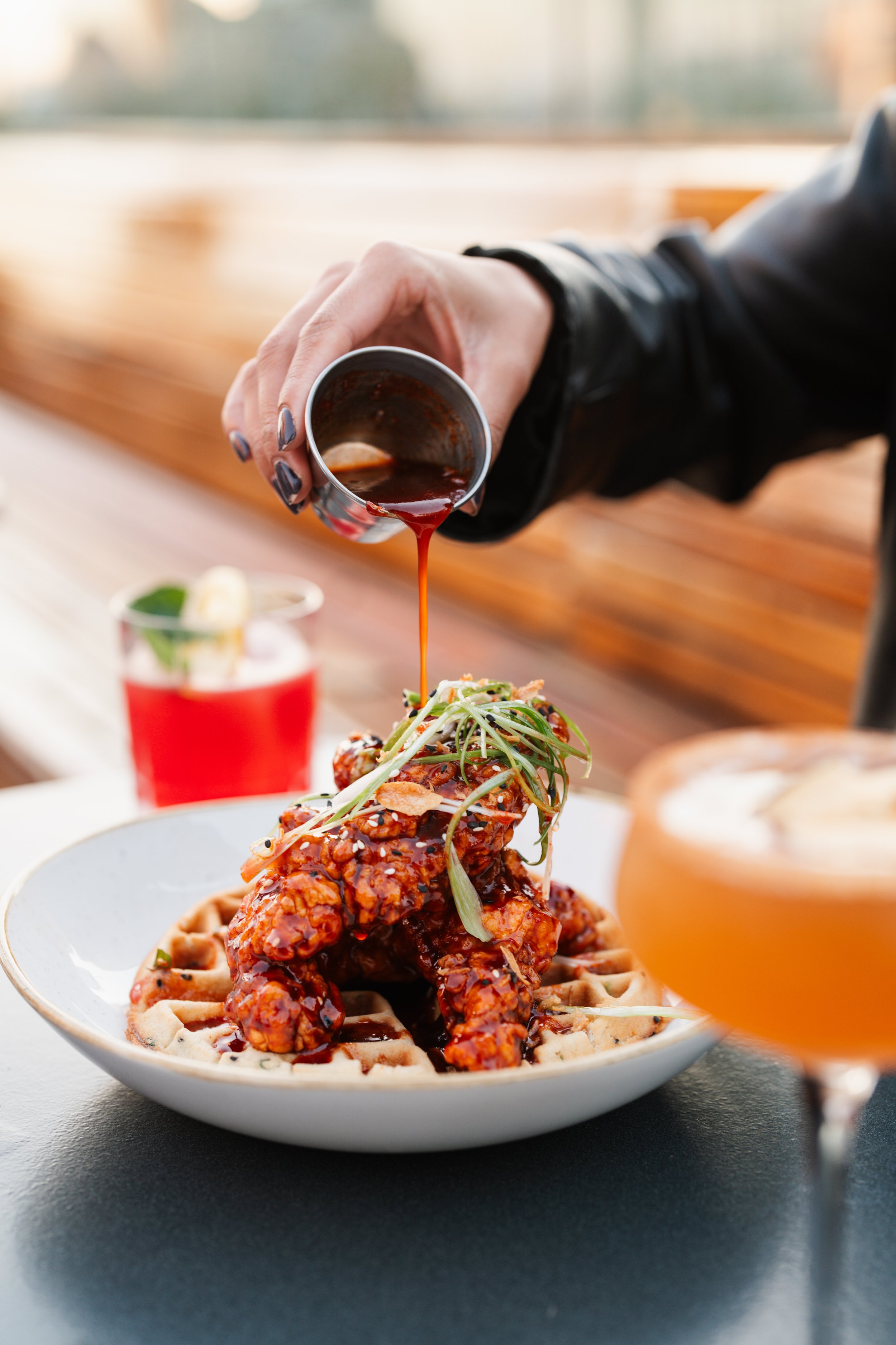 Person pouring sauce over a plate of chicken and waffles with garnish, near various drinks.