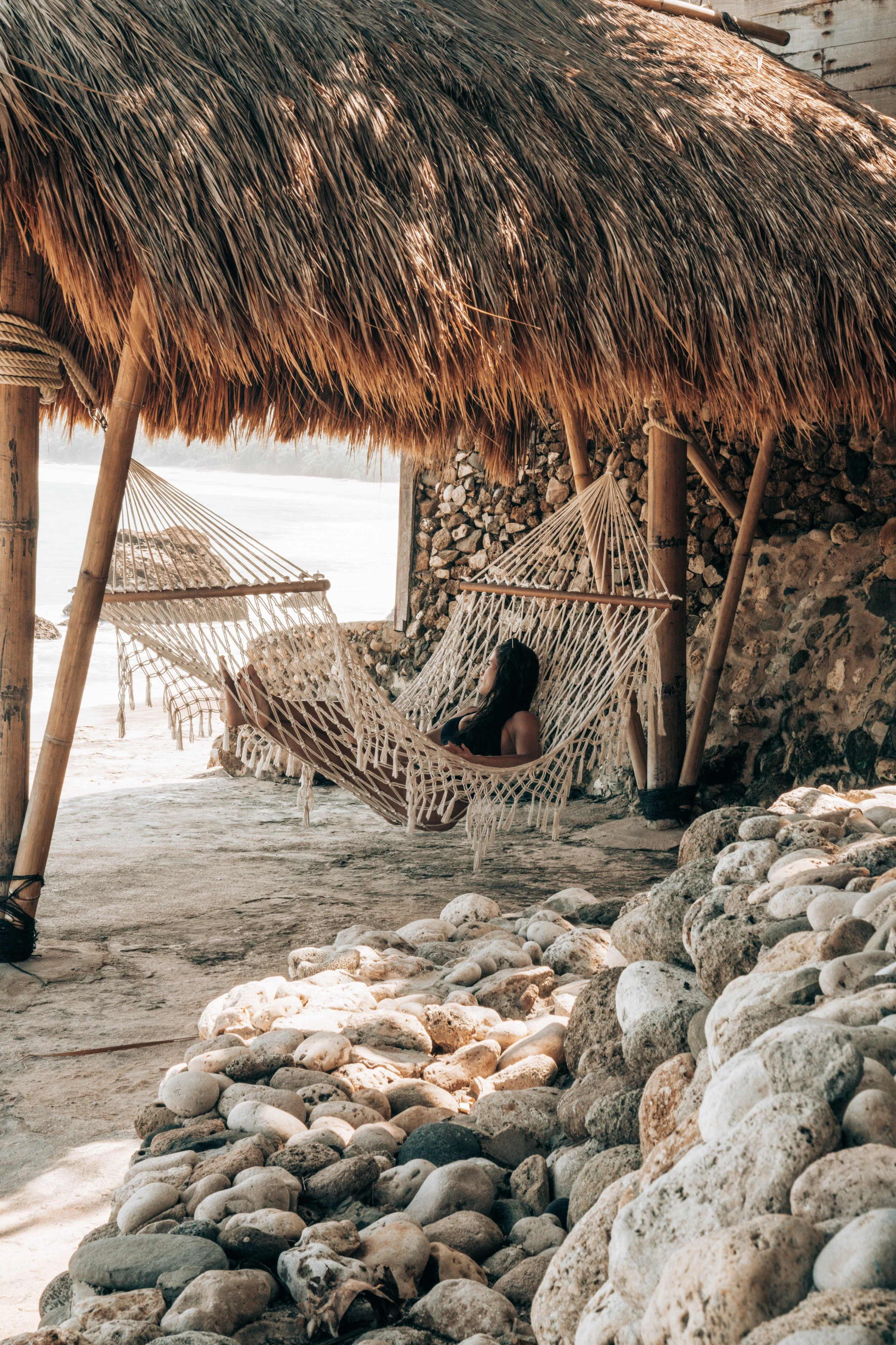 A woman relaxing on a hammock under a thatched roof on a beach with rocks and a view of the ocean in the background.