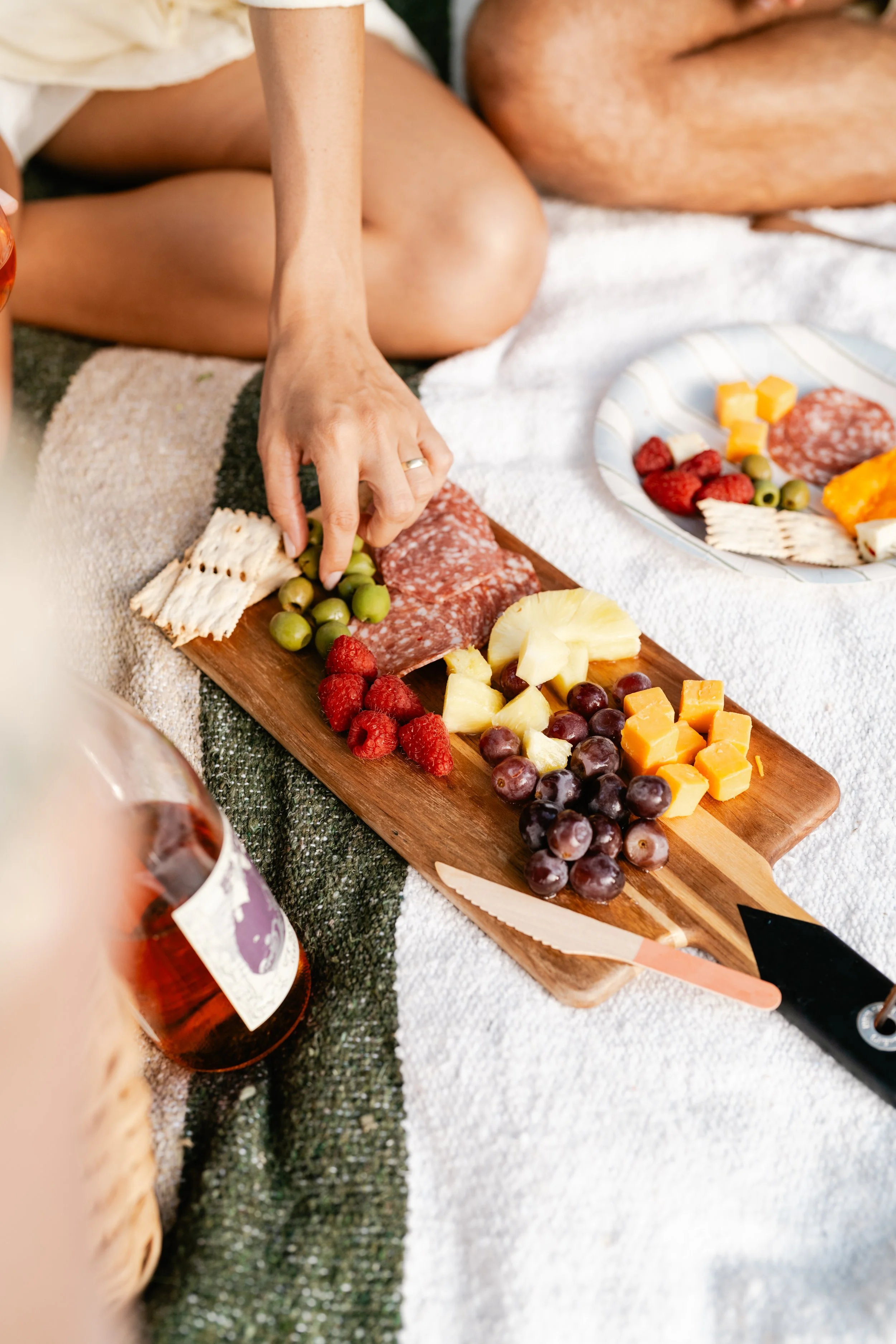 Close-up of a person reaching for grapes on a wooden cheese board, with cheese, raspberries, pineapple, grapes, and salami, on a picnic blanket.