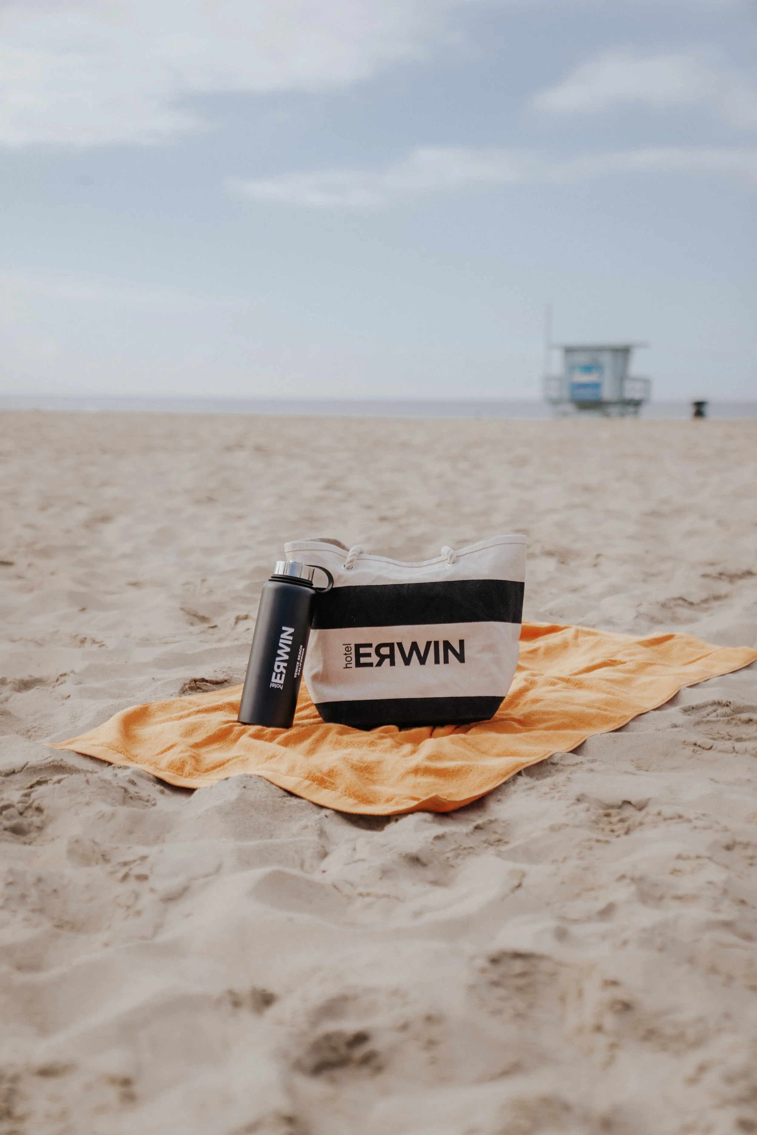A beach scene with an orange towel on the sand, a black water bottle, and a striped tote bag with the words 'hotel ERWIN' on it. In the background, a lifeguard tower is visible against a partly cloudy sky.