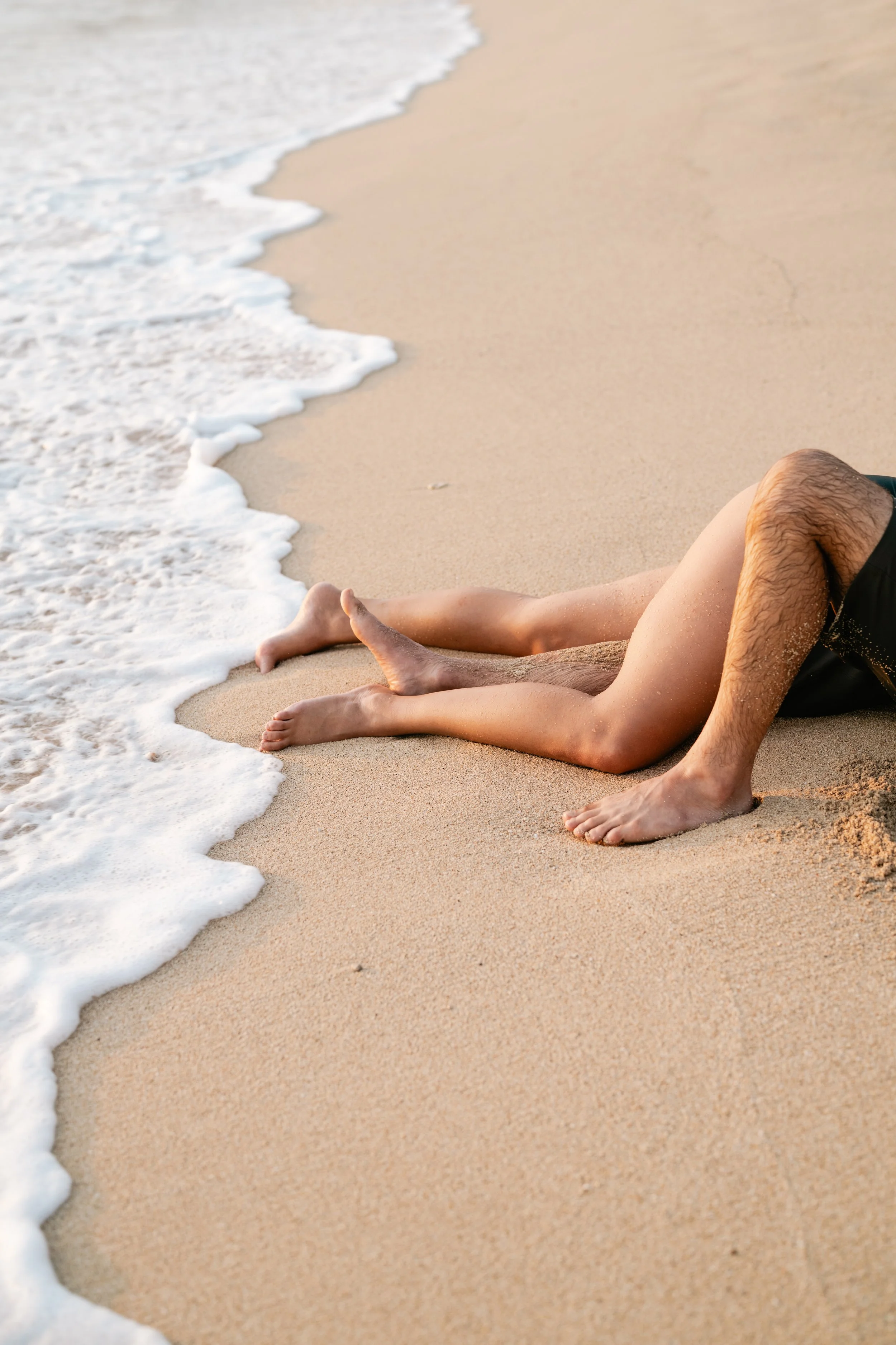 Two people lying on a sandy beach near the shoreline with waves washing over their legs.