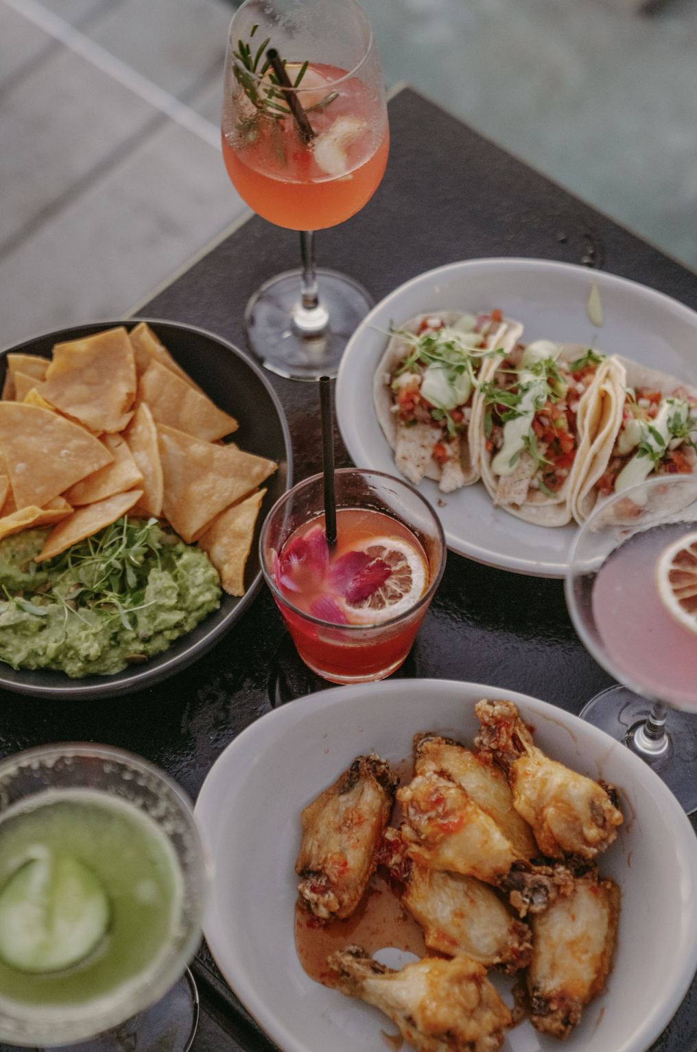 Assorted Mexican dishes on a black table including guacamole with tortilla chips, tacos topped with cilantro and sauce, fried chicken wings, and two colorful cocktails with fruit garnishes.