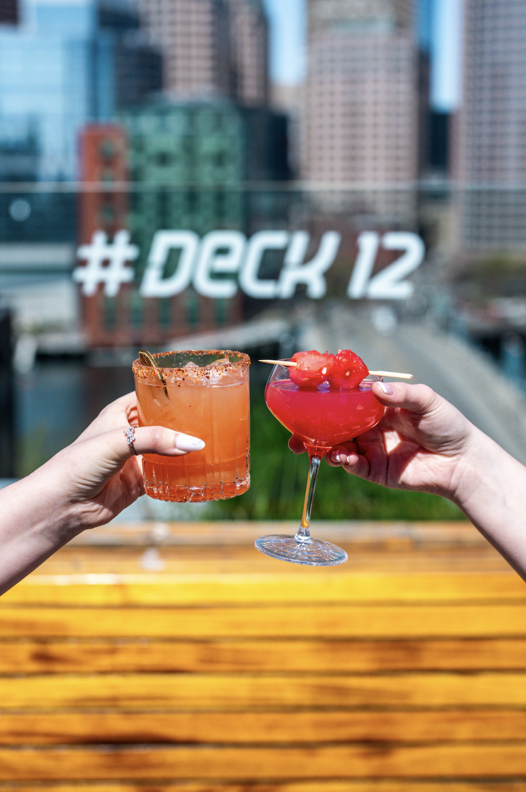 Two hands holding cocktails with a backdrop of city buildings and a glass window with the hashtag '#DECK 12' written on it.