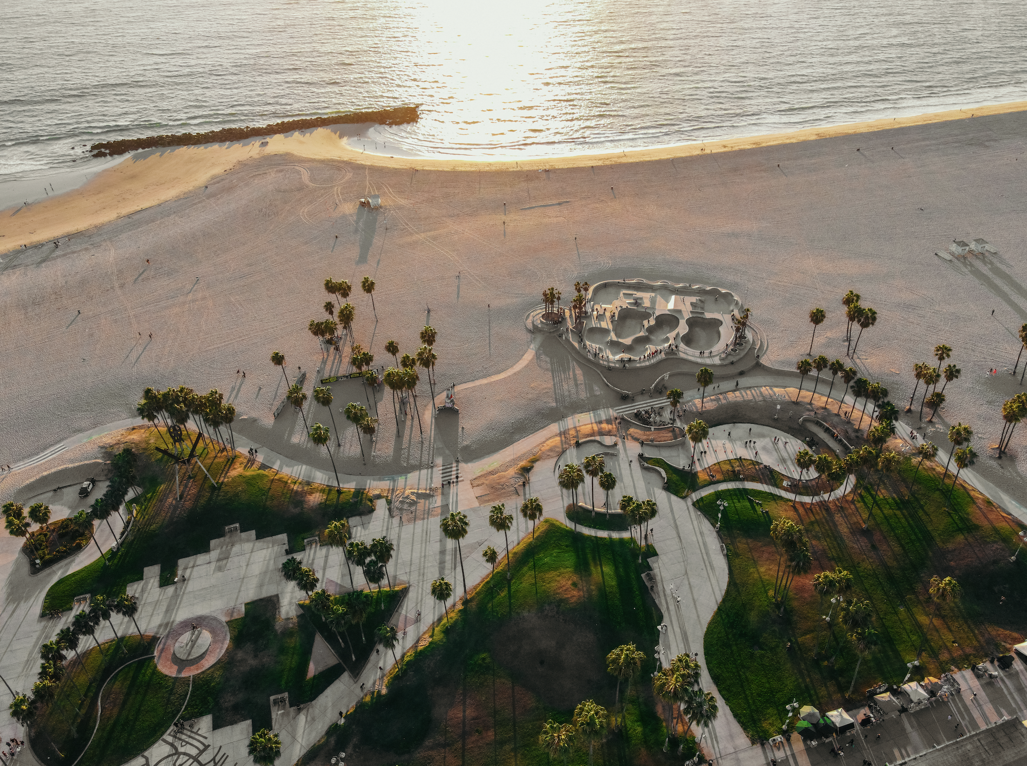 An aerial view of a beachfront park during sunset, with palm trees, pathways, and a skate park, near the ocean shoreline.
