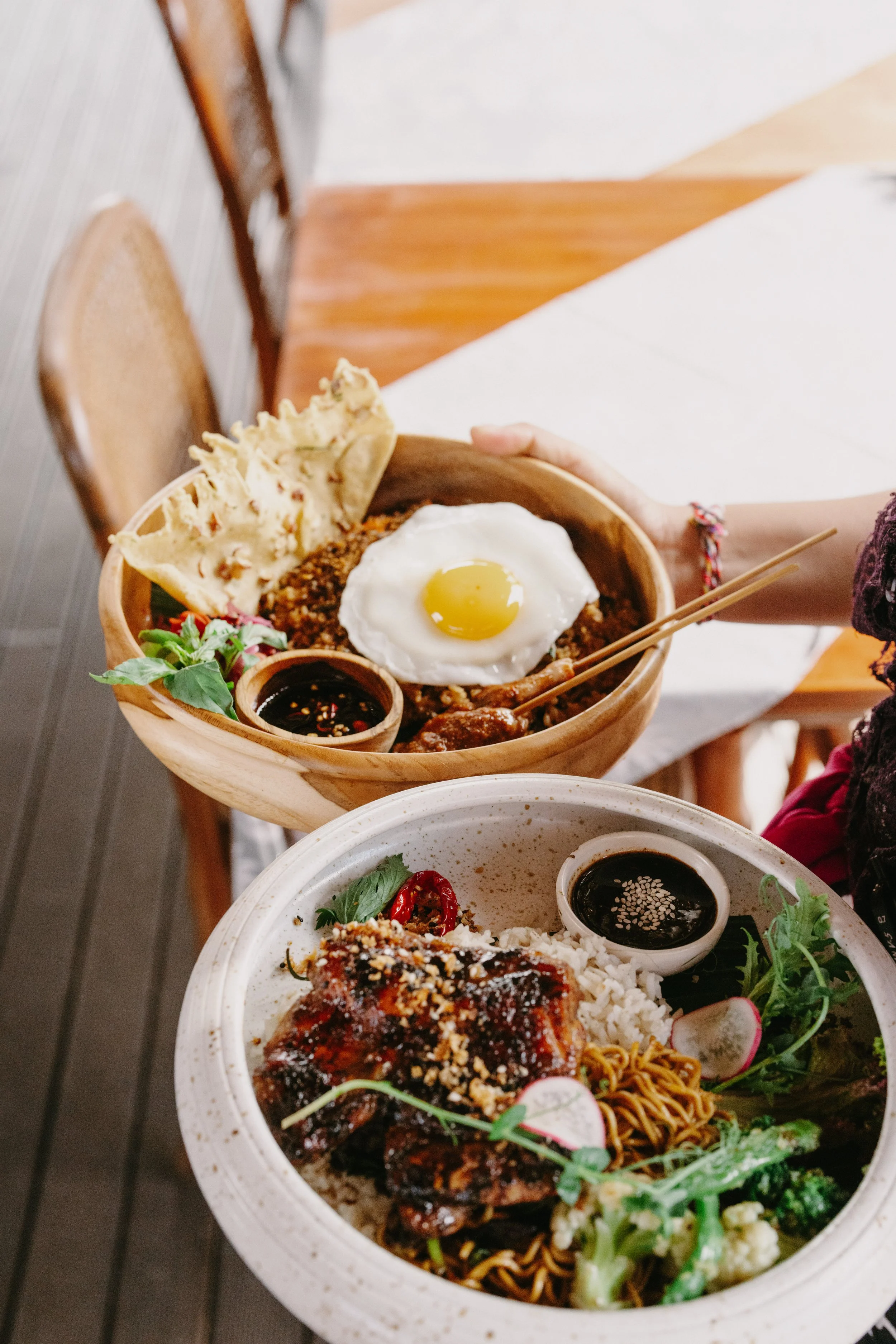 Two bowls of Asian-inspired dishes, one with rice, grilled chicken, and vegetables, and the other with rice, ground meat, a fried egg, and dipping sauces.