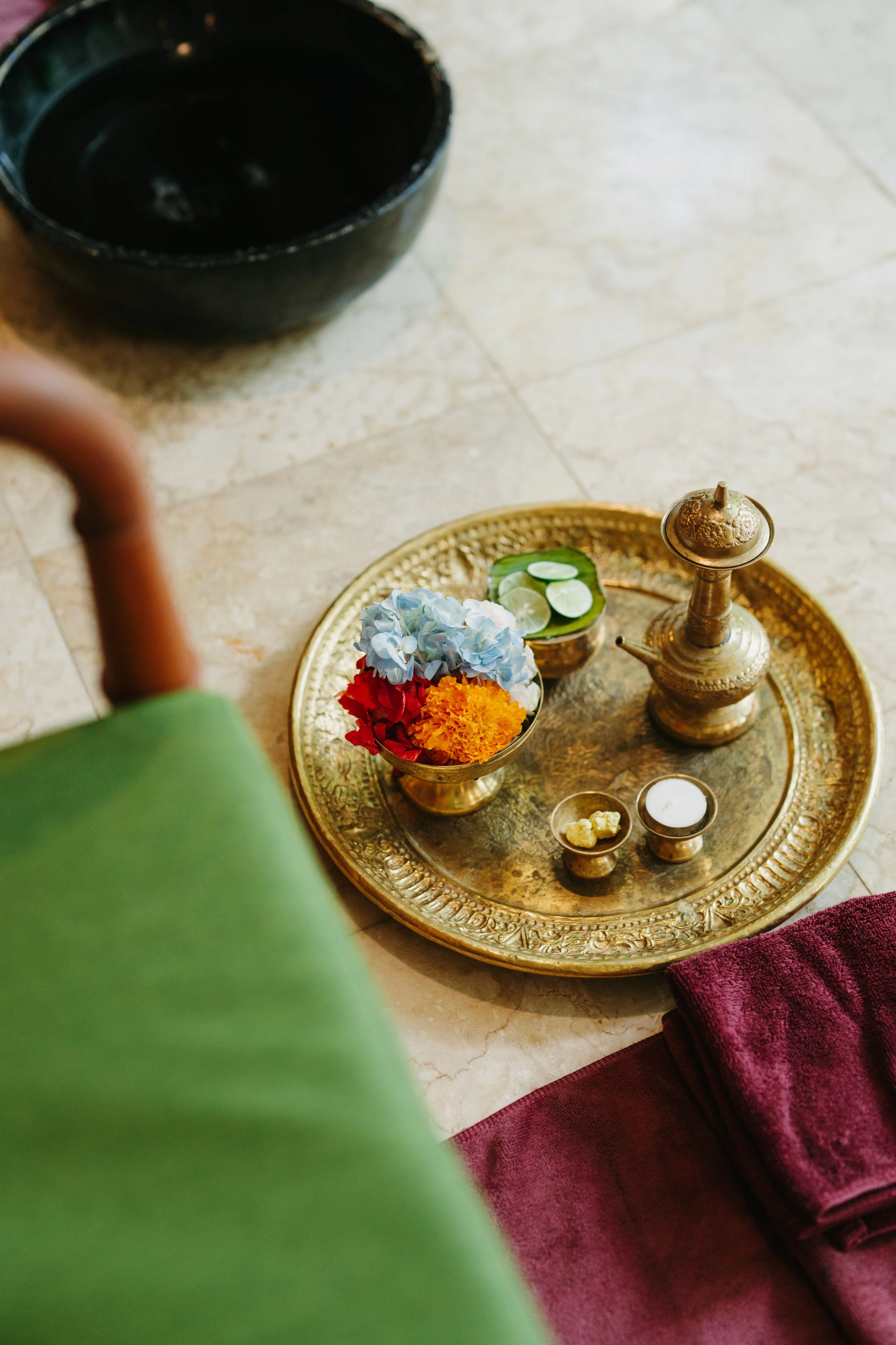 A brass tray with traditional Indian decorative items, including an ornate brass vessel, small bowls with flowers, lime slices, and a candle, is placed on the floor next to a folded maroon towel, with a black bowl and a green chair partially visible 