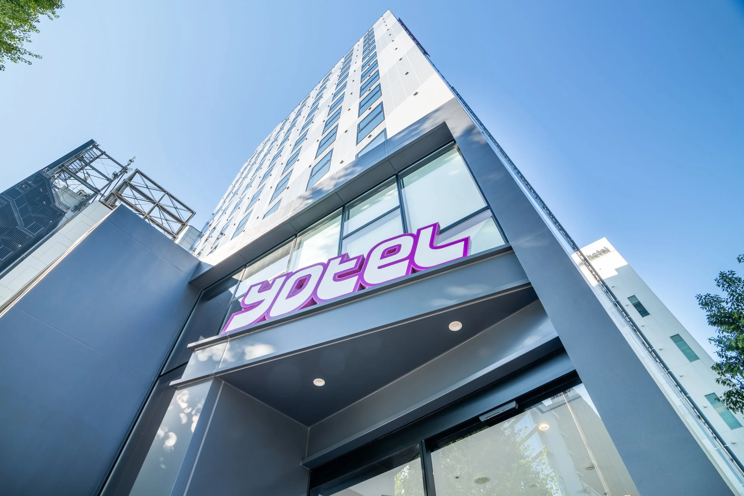 Low-angle view of a modern hotel building with a purple sign that says 'hotel' at the entrance, tall skyscrapers and blue sky in the background.