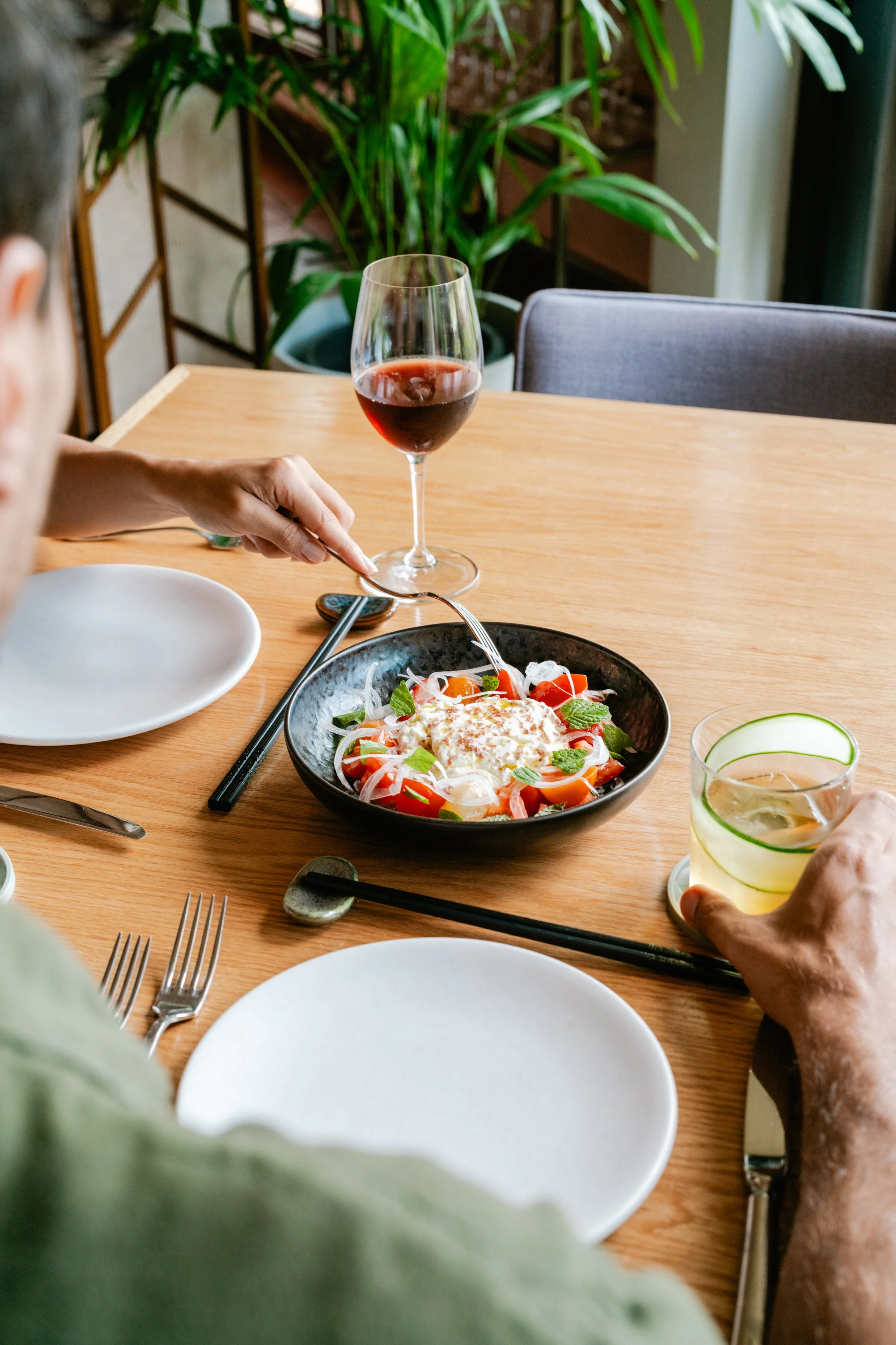 A dining table with a black bowl of salad topped with cheese, surrounded by a glass of red wine, a glass of water with cucumber slices, and various utensils and empty plates.
