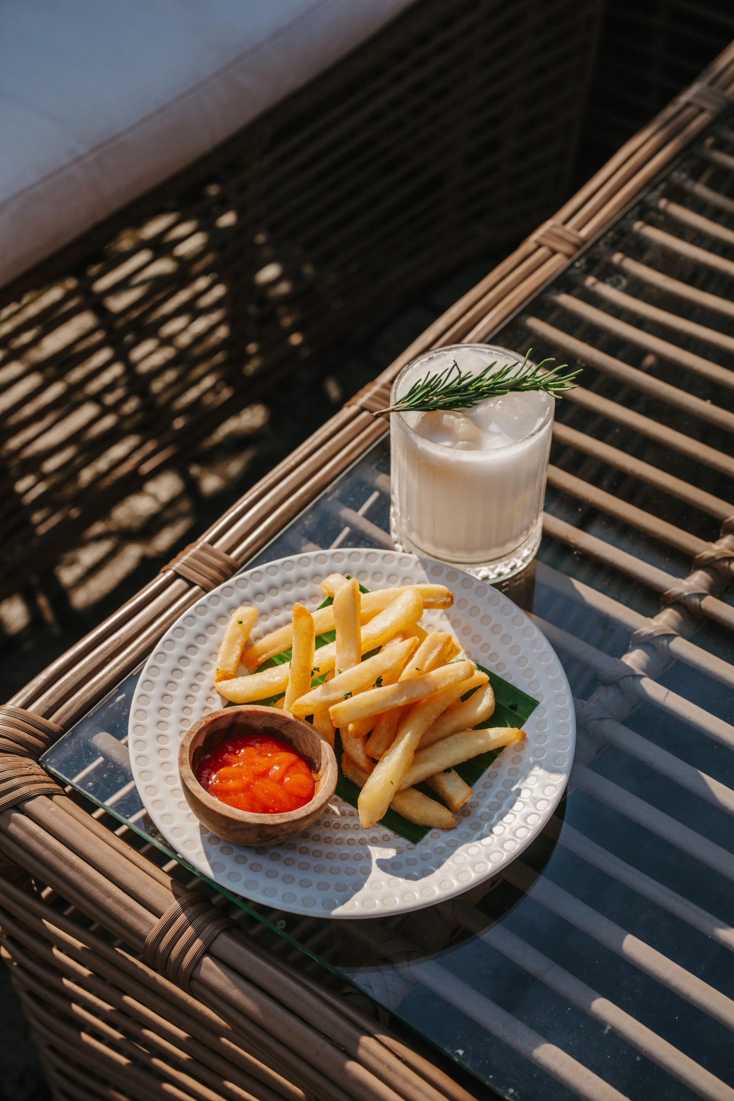 Fried French fries with a small bowl of ketchup on a white plate, a glass of cream drink garnished with a sprig of rosemary, on a glass-topped wicker table with sunlight and shadows.