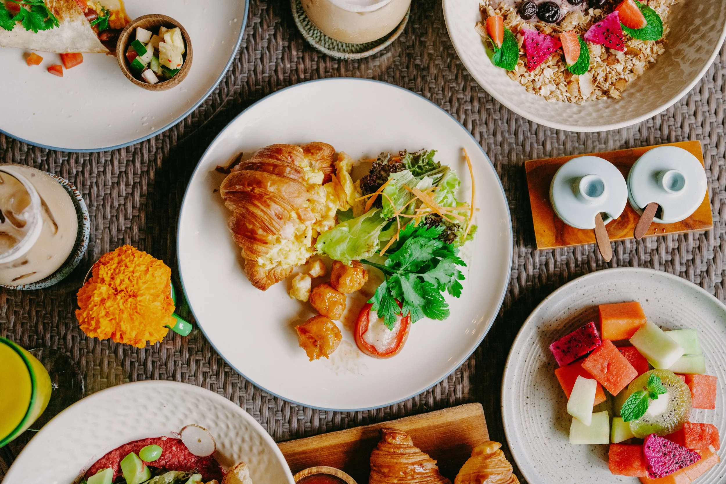 A top-down view of a breakfast spread including a croissant with scrambled eggs and salad, bowls of fruit salad and granola with strawberries, blueberries, and mint, a glass of iced coffee, small containers of milk or cream, and a marigold flower on 