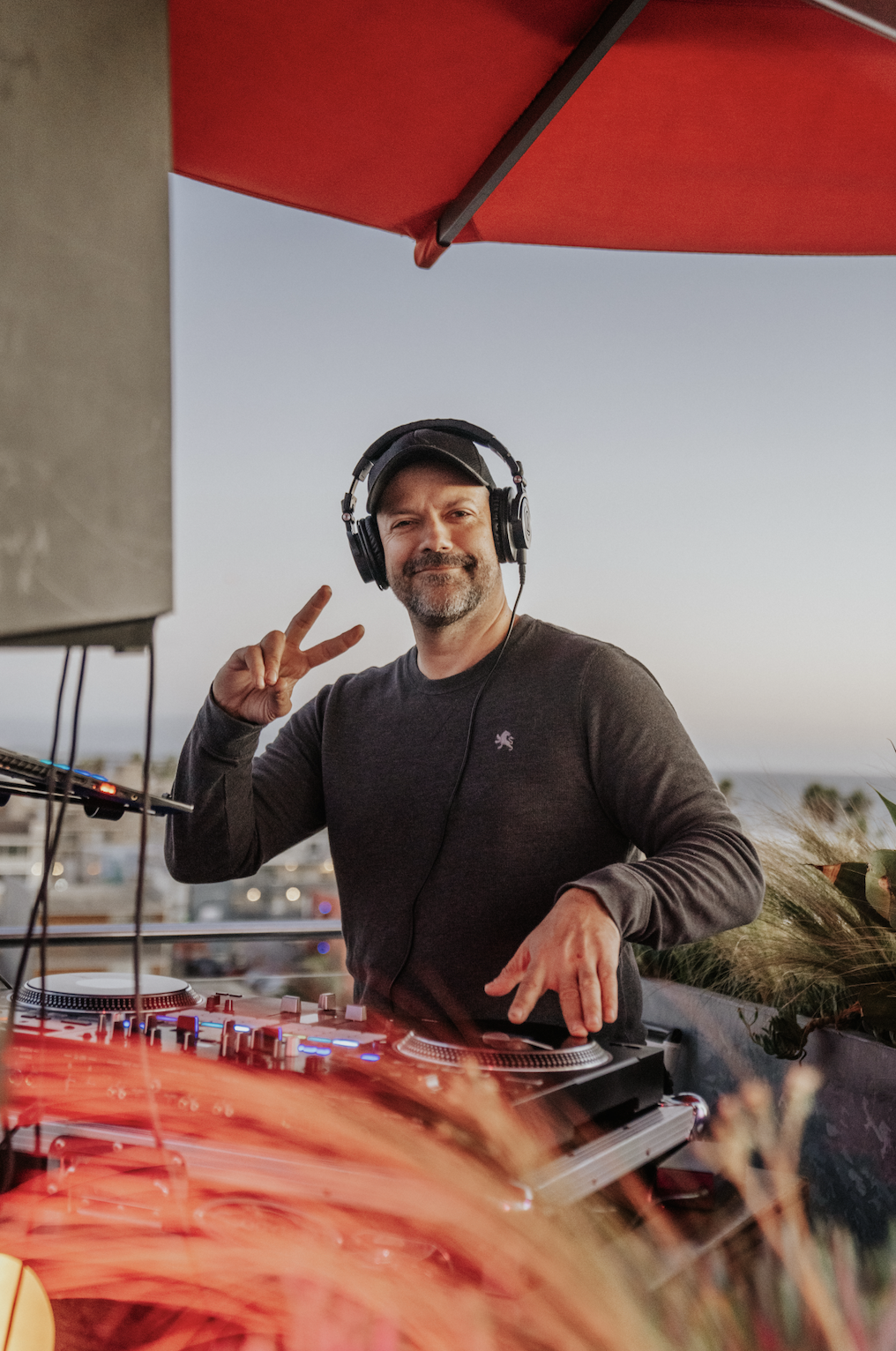 A man wearing headphones and a dark long-sleeve shirt is DJing, smiling, and making a peace sign with his right hand while standing outdoors at sunset under a red umbrella.