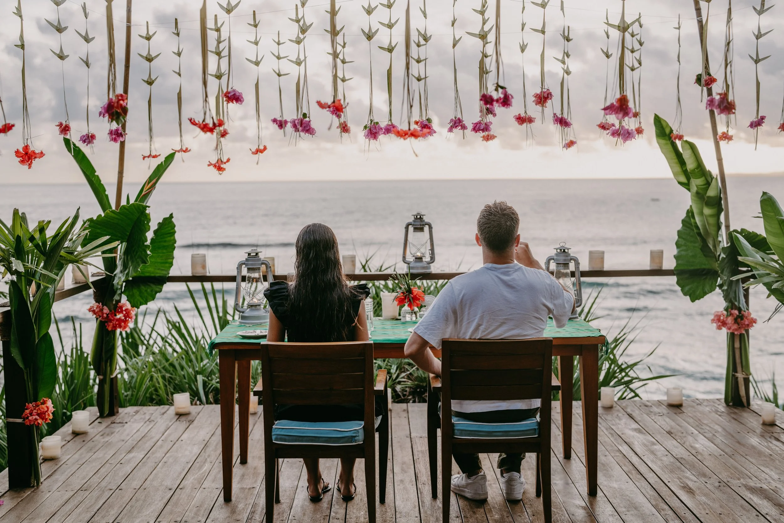 A couple sitting at a table on a wooden deck overlooking the ocean, decorated with pink and red flowers hanging from above and plants on the sides, with lanterns and candles on the table.