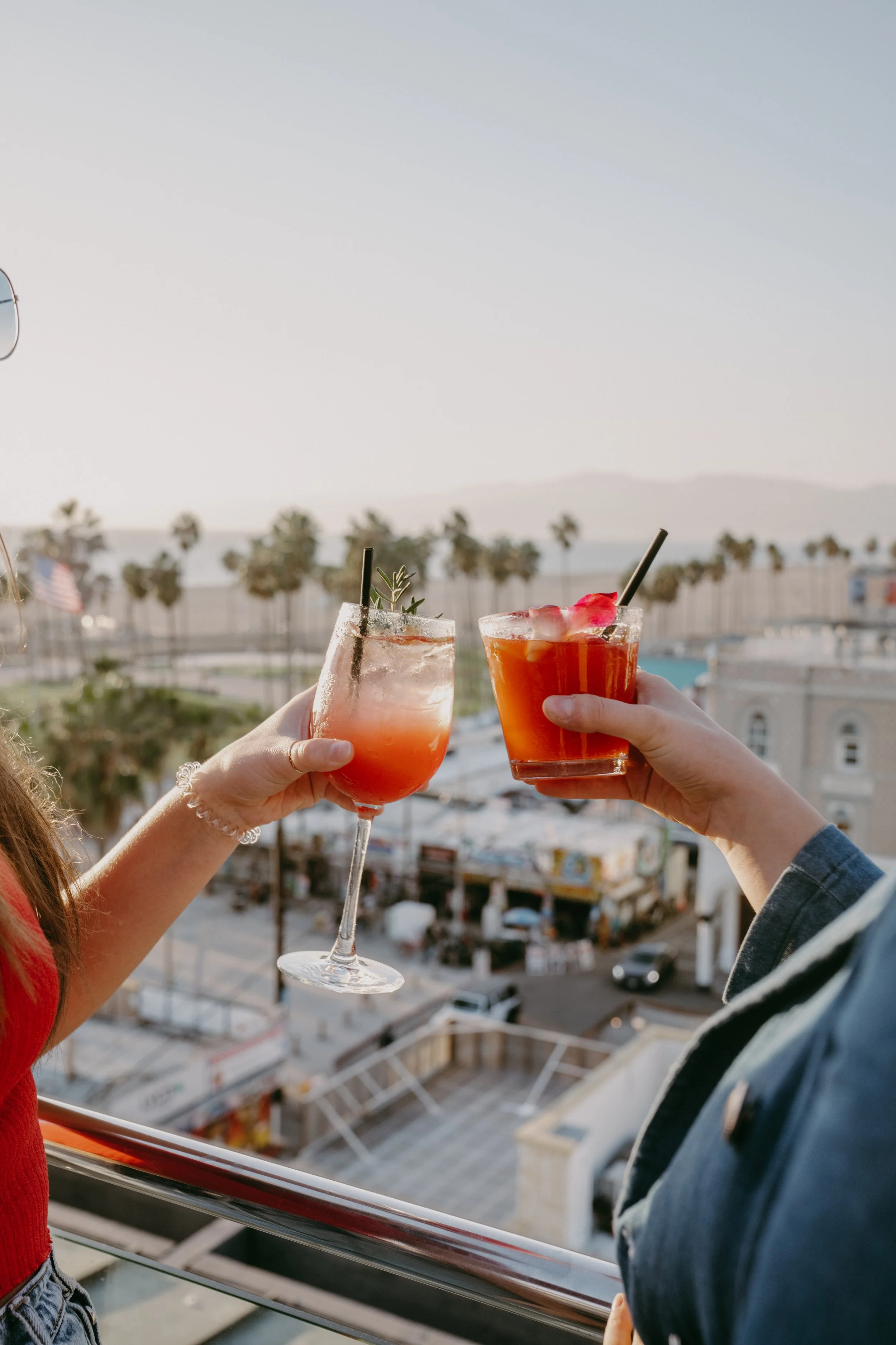 Two people holding drinks in outdoor setting with a view of palm trees and cityscape at sunset.
