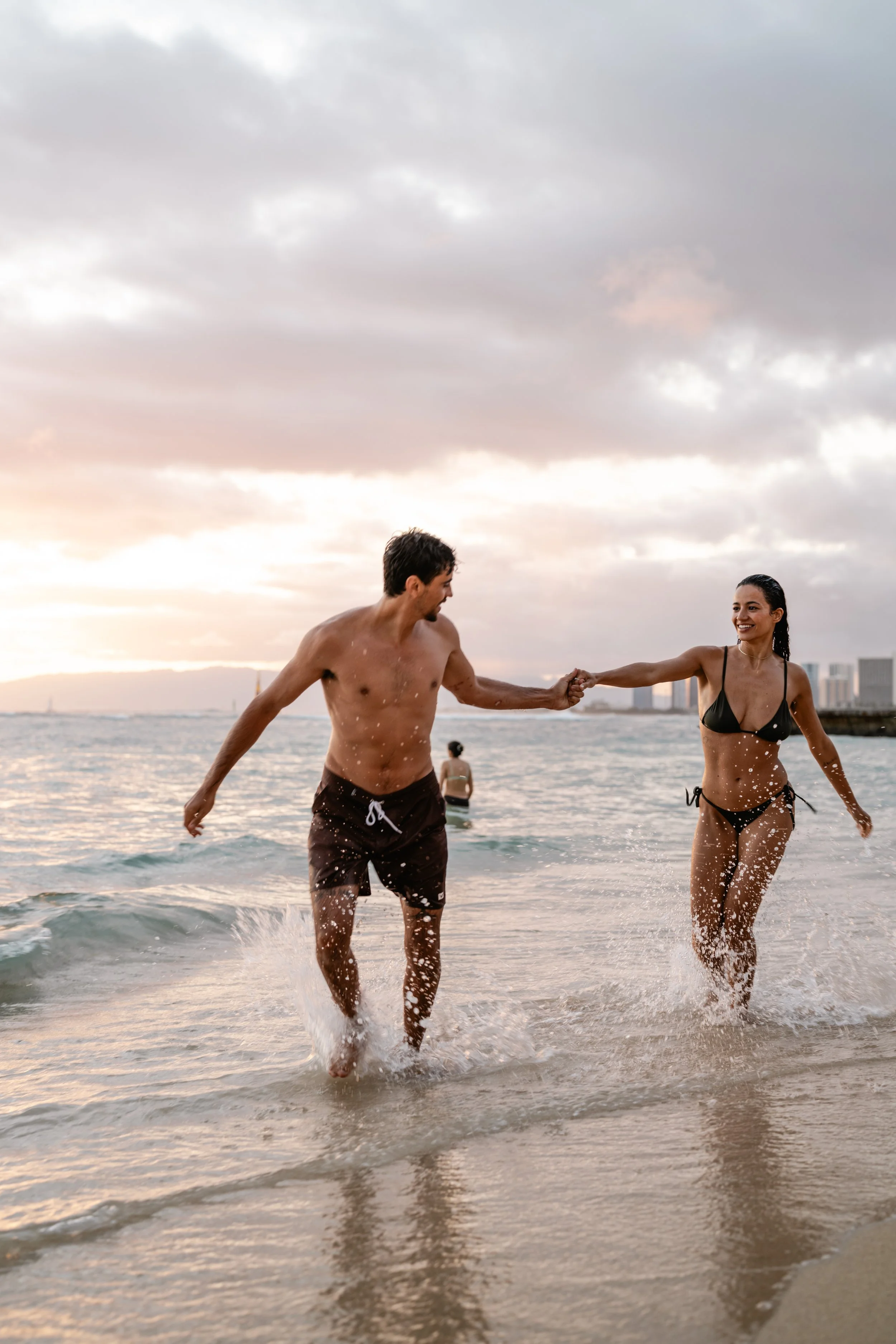 A man and woman in swimsuits playing and holding hands in the shallow water at the beach during sunset.