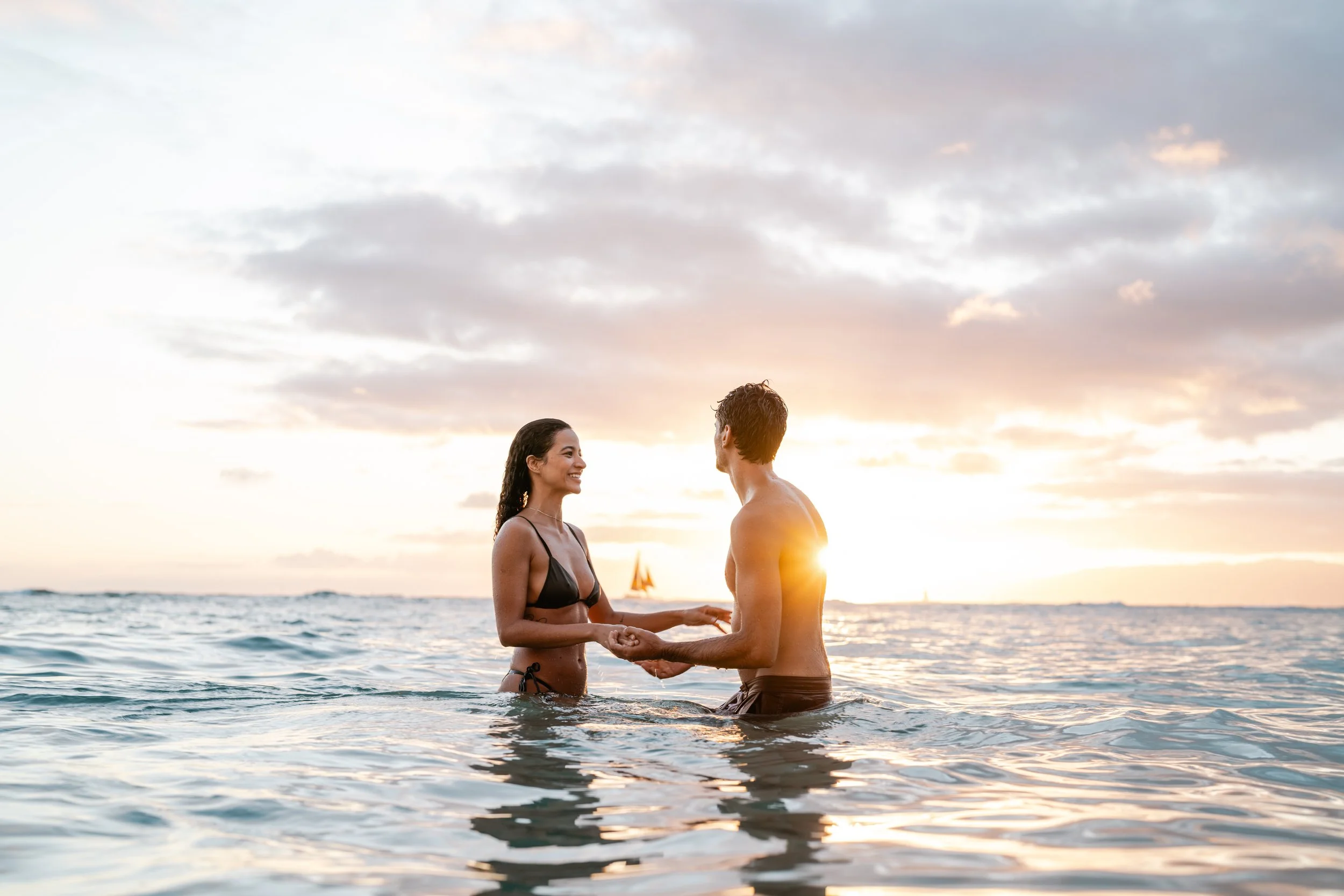 A couple holding hands and smiling at each other while standing in the ocean at sunset, with a sailboat in the background.