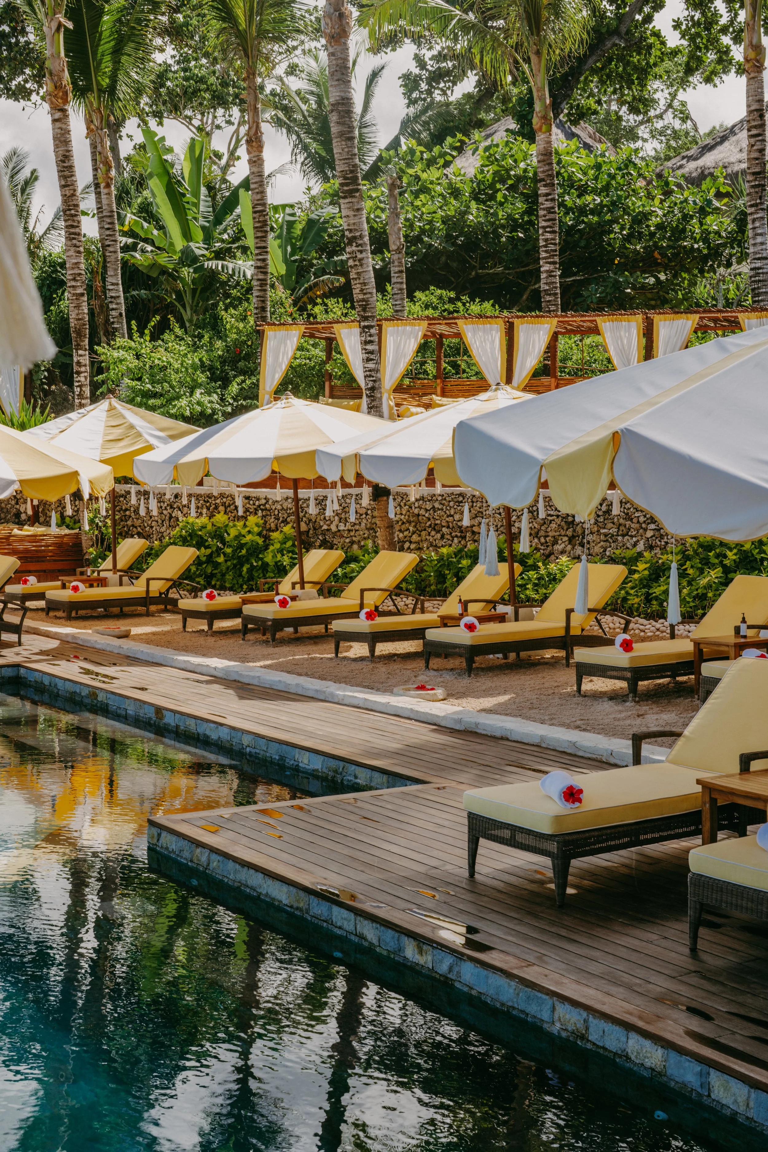 Poolside area at a tropical resort with yellow lounge chairs under white umbrellas, decorated with flowers, and surrounded by green palm trees and lush vegetation.