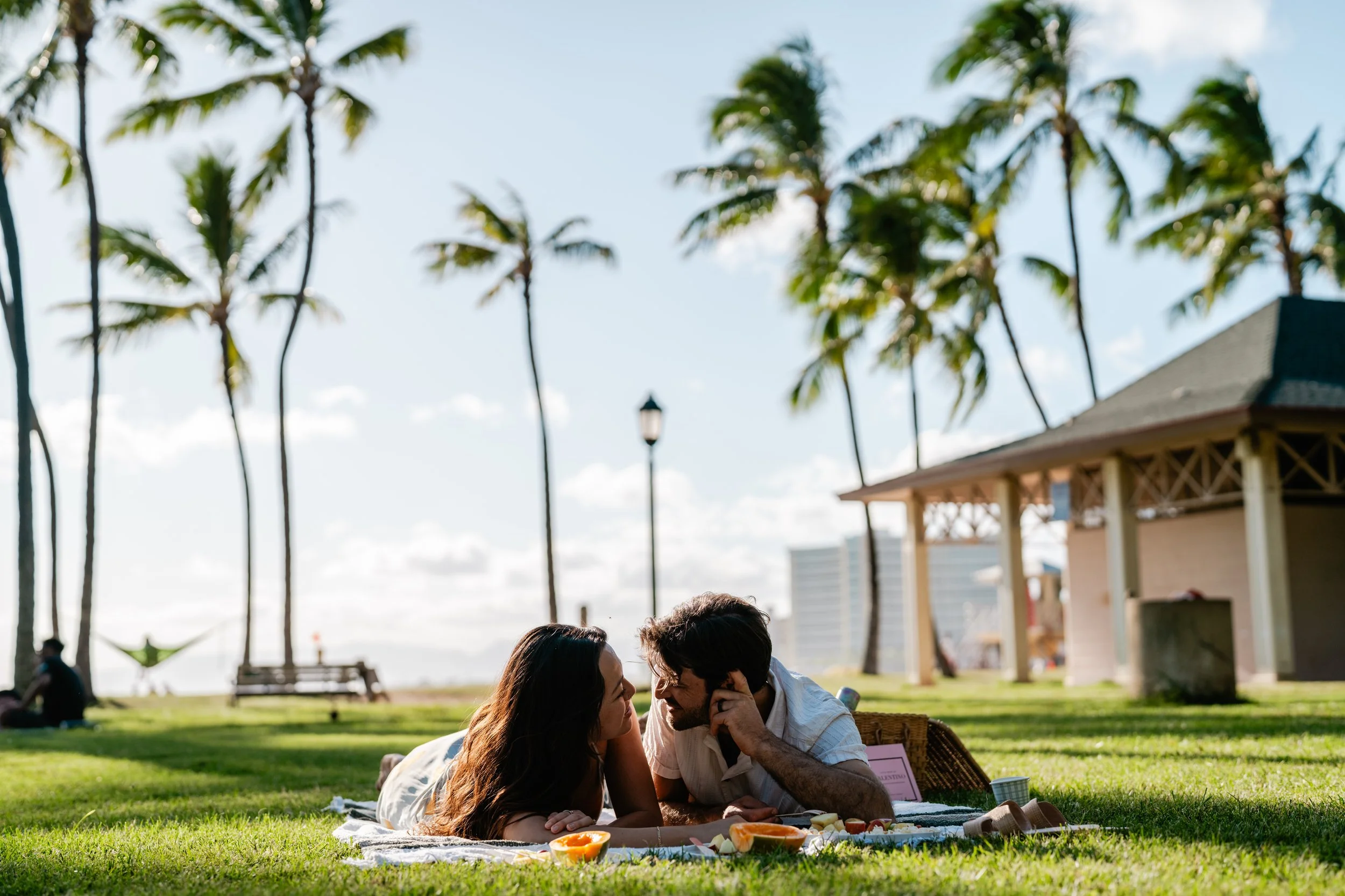 A couple having a picnic on the grass near the beach with palm trees in the background.