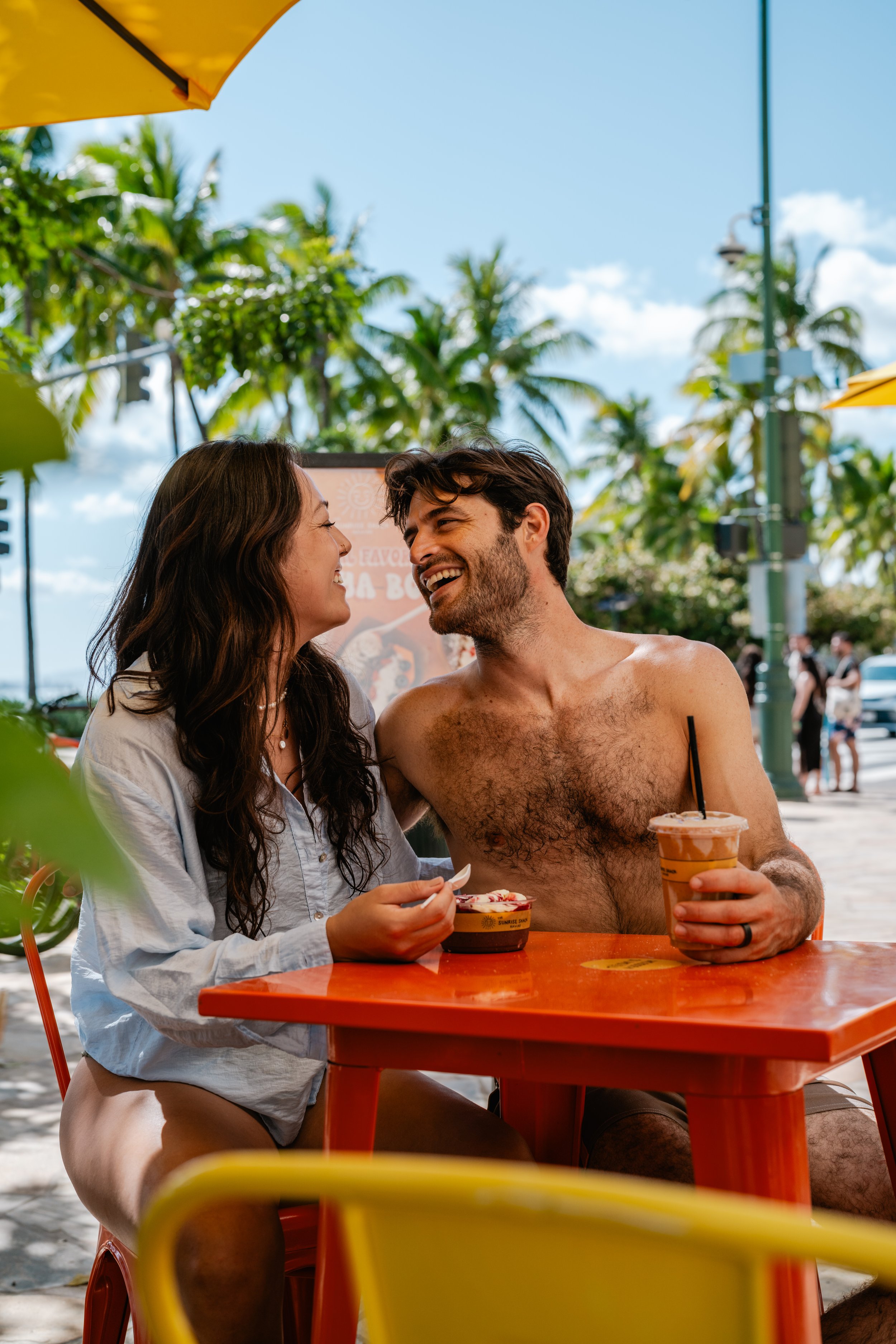 A couple sitting at an outdoor table on the beach, smiling at each other, with palm trees and a blue sky in the background. The woman has long brown hair and is holding food, while the man, shirtless, is holding a drink.