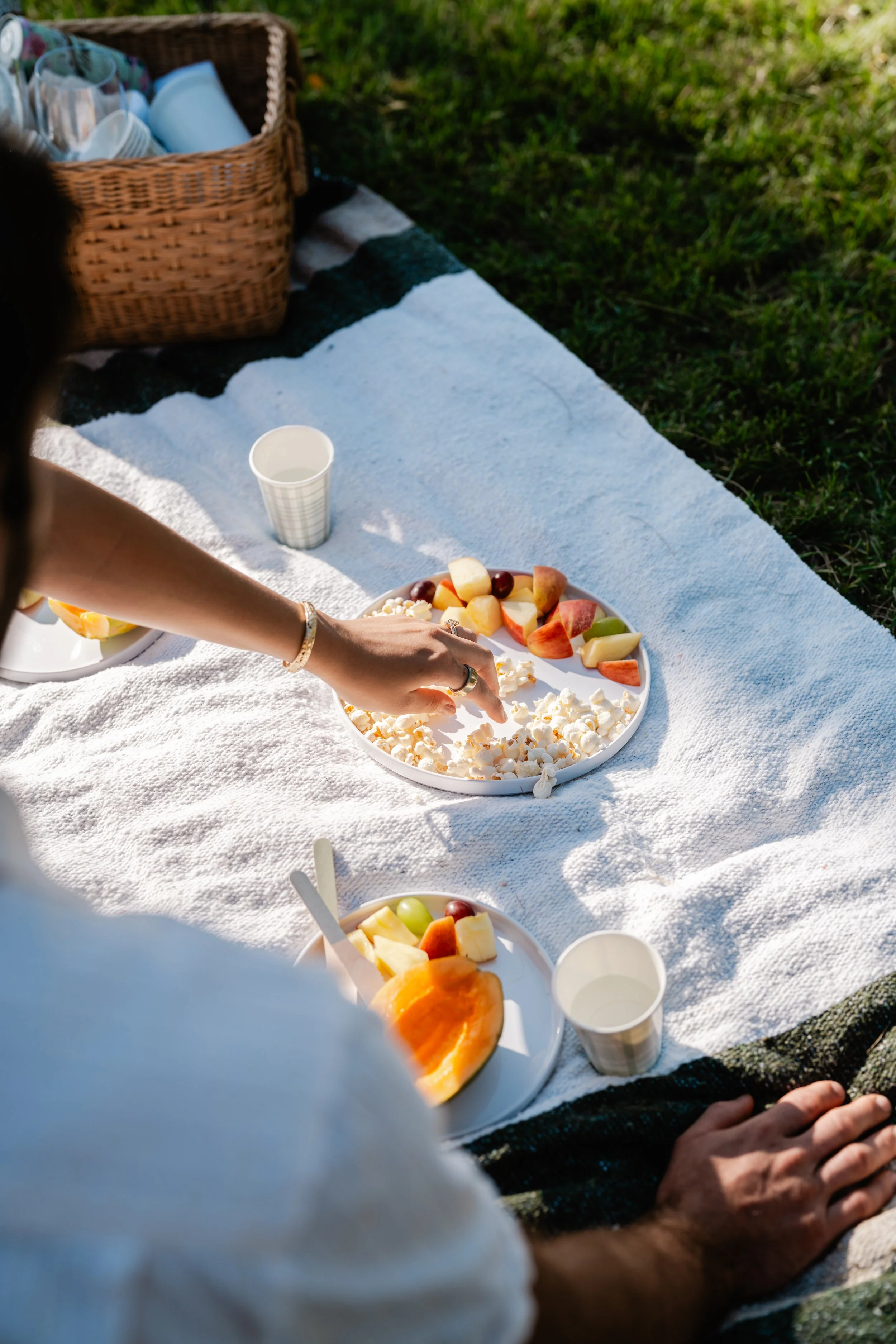 A picnic setup on a white blanket outdoors with plates of sliced fruit, cheese, and popcorn, along with cups and a basket of food.