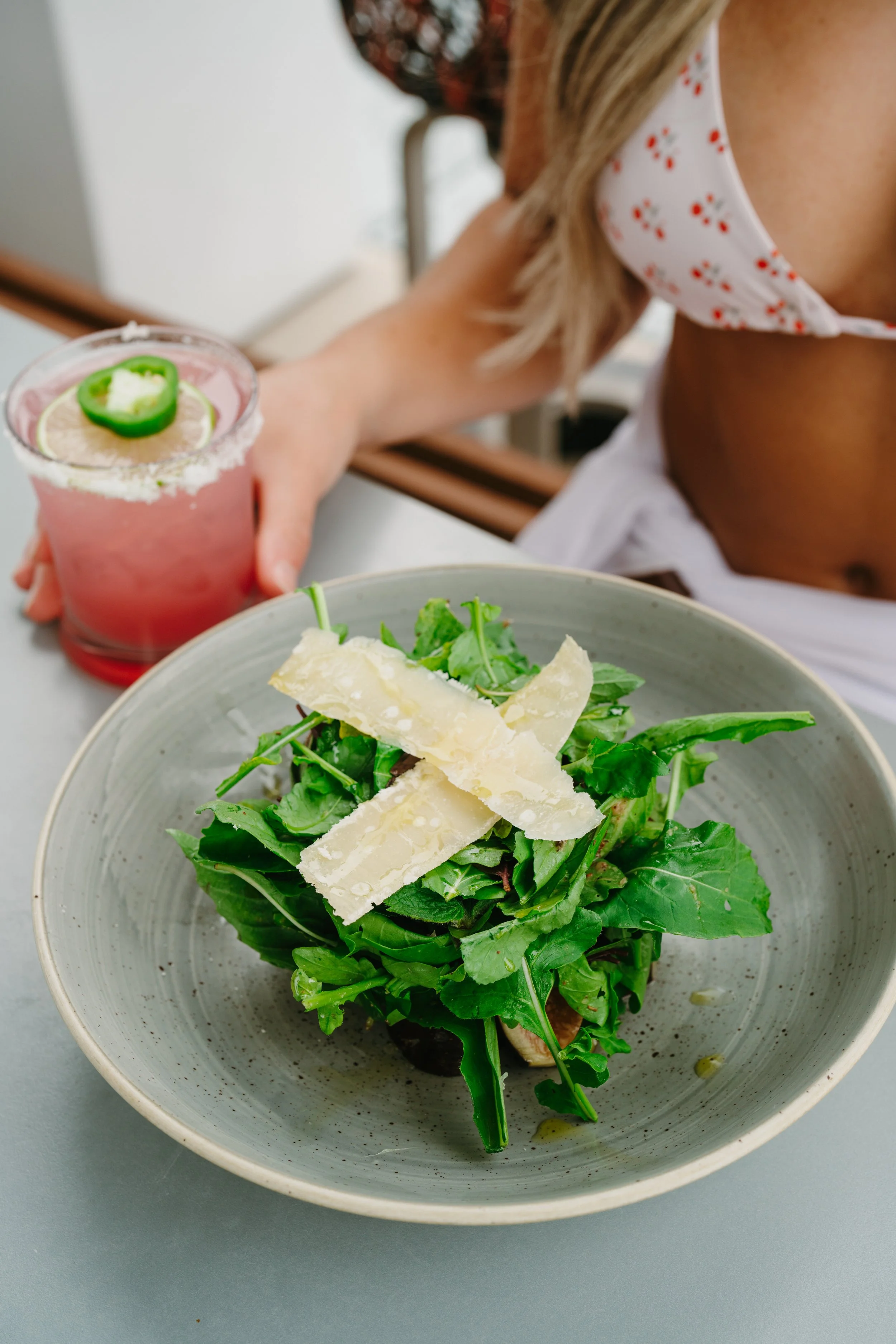 A bowl of salad topped with cheese shavings, accompanied by a pink cocktail drink garnished with a green pepper slice and lemon slices. A woman wearing a floral face mask is reaching towards the drink.