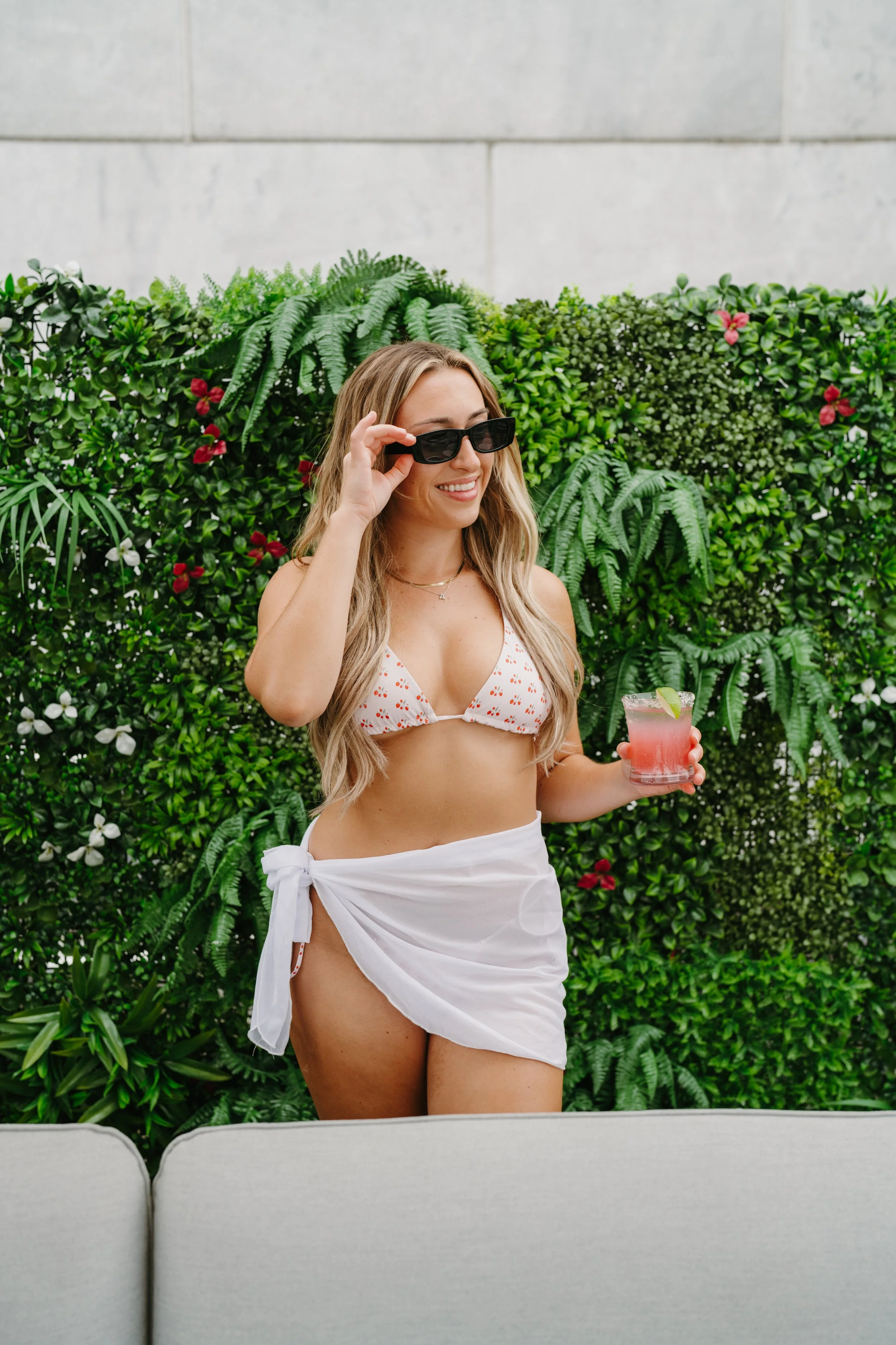 A woman in a bikini top and white sarong holding a pink cocktail while adjusting her sunglasses against a lush green leafy background.