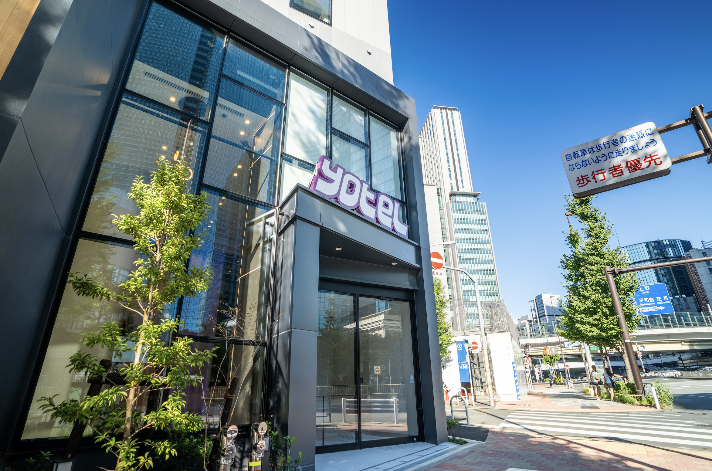 Modern building entrance with a sign that says 'YELP' and a tree in front, with bright blue sky and tall skyscrapers in the background.