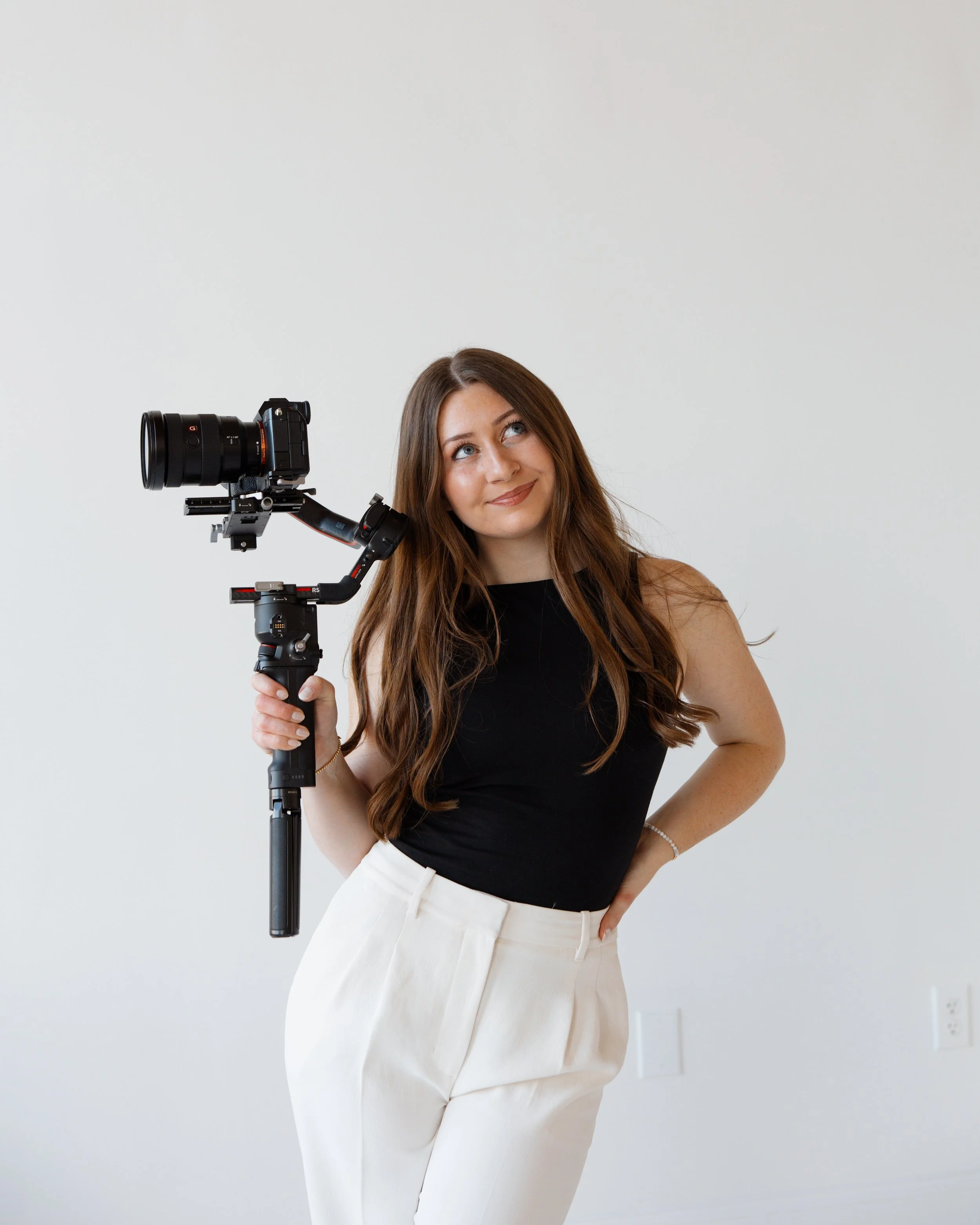 Videographer + Photographer, Young woman with long brown hair holding a camera stabilizer, smiling and looking up.