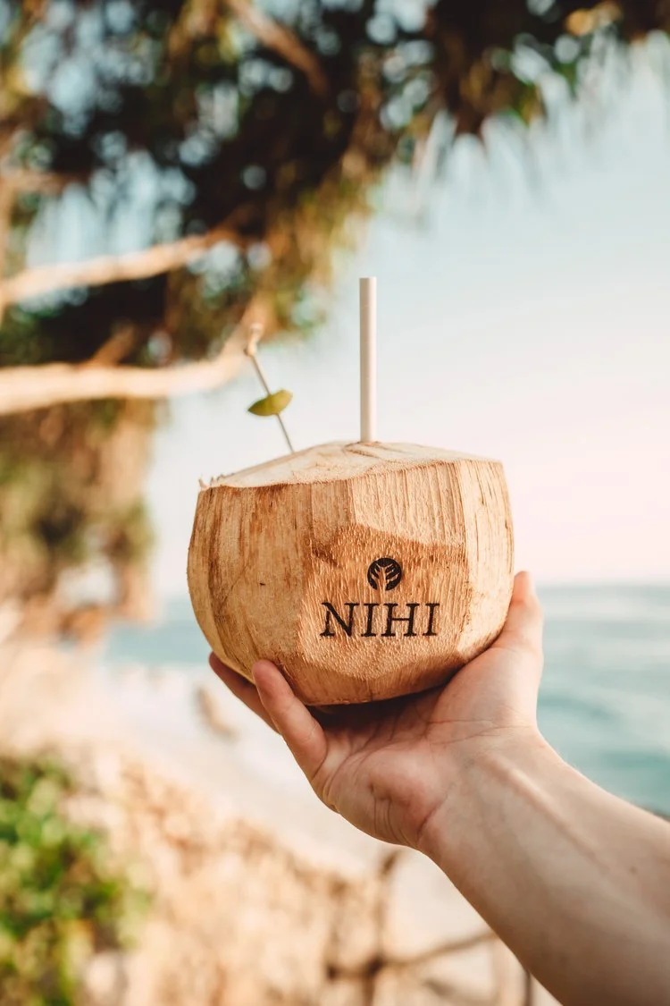 Person holding a young coconut with a straw and a small yellow umbrella, with a beach and tree in the background.