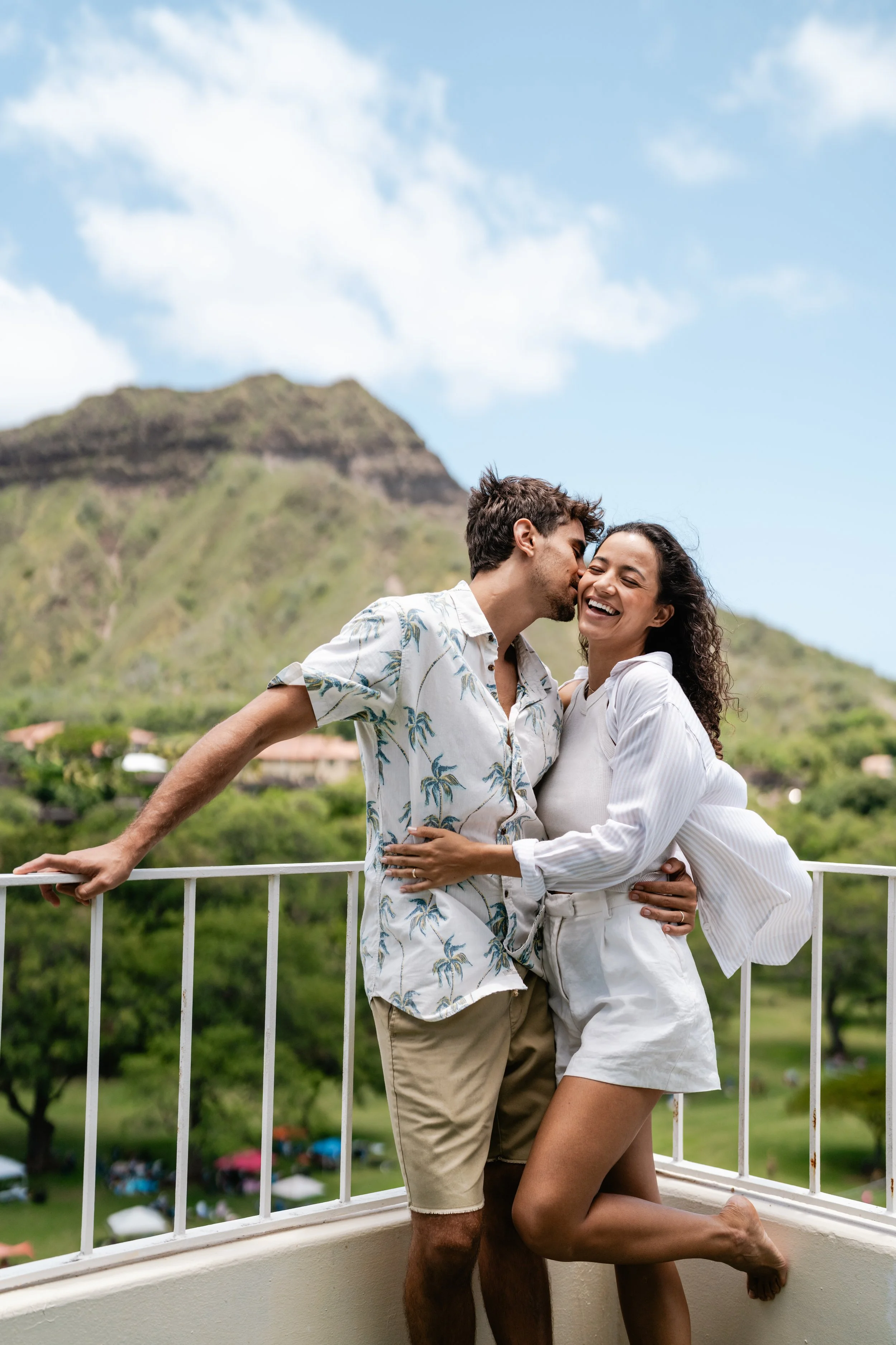 Hotel & Resort Photography & Videography. A happy couple embraces on a balcony with a mountain and greenery in the background. The man is whispering into the woman's ear as she laughs.