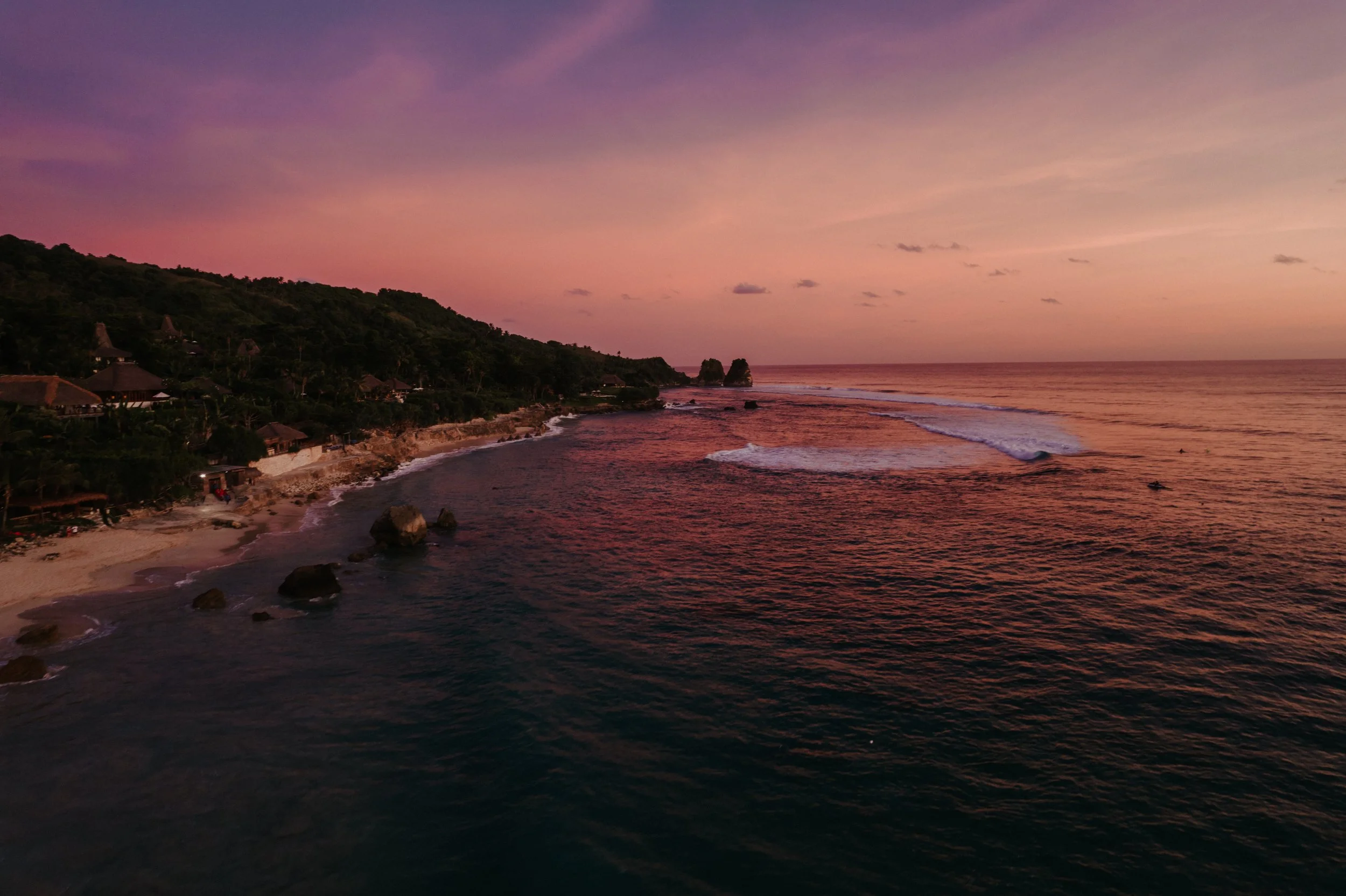 Sunset over a tropical beach with waves crashing and a green hillside with small huts lining the shore.